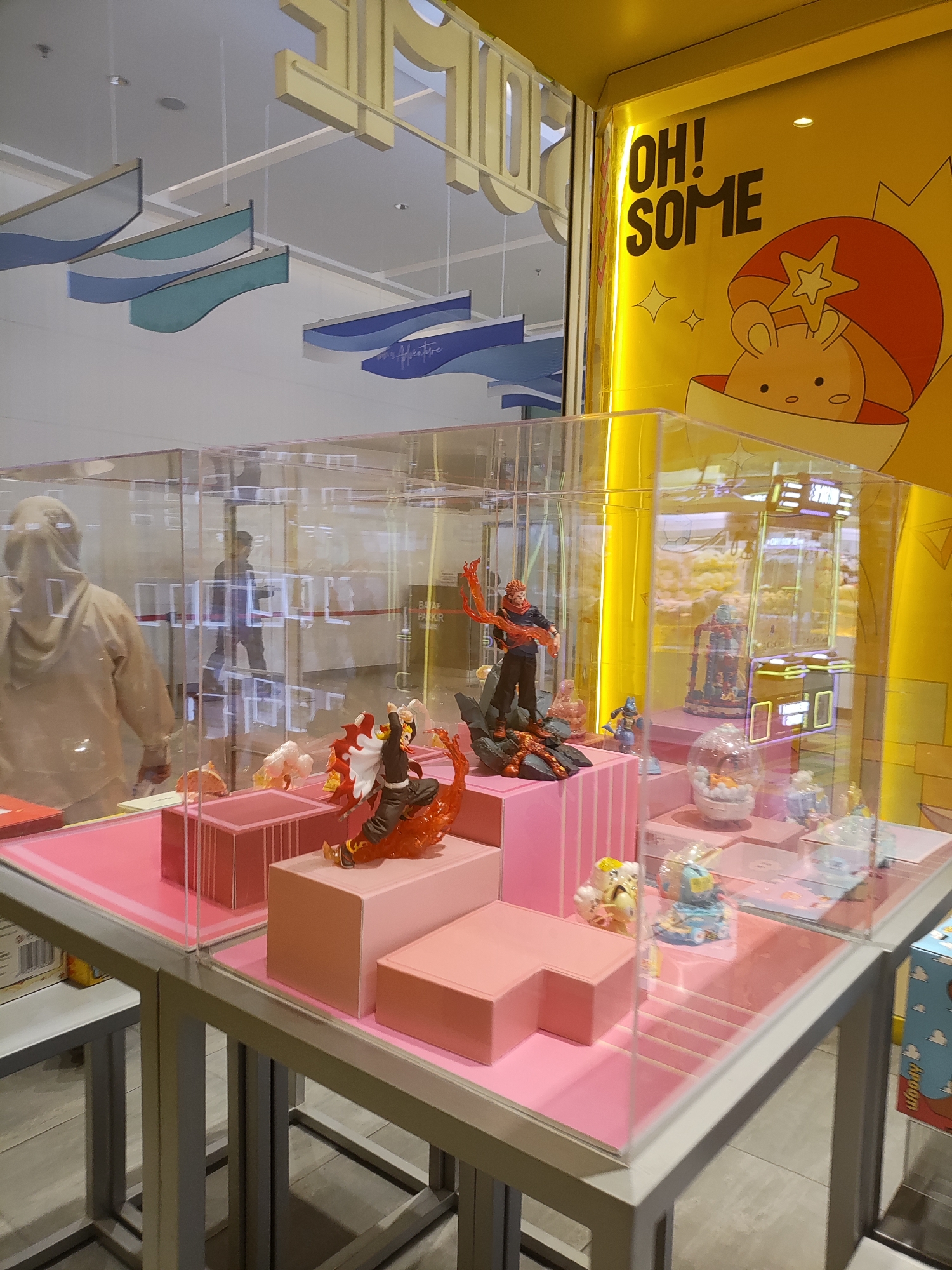 The image depicts an indoor retail display, likely a toy store or a section of a larger store, with a strong emphasis on anime-style figurines. The display is enclosed in clear acrylic cases, revealing a collection of collectible figures arranged on pink and white tiered platforms. In the foreground, a figure with fiery effects and a character in a black outfit with red accents are prominent. Other smaller figurines and decorative elements are visible within the cases.
In the background, the store features a bright yellow wall with a stylized illustration and text that reads "OH! SOME." The ceiling has a series of decorative, wave-like panels in blue and green hues. Through the acrylic, another person is visible in the background, walking with their back to the camera. To the left, a person wearing a tan hooded garment stands facing away, partially obscured by the display case.
The overall environment is clean and well-lit, typical of a modern retail setting. The presence of specific characters suggests a focus on popular anime or manga franchises.
This image was taken inside the XXI Cineplex at the Summarecon Mall Bekasi, located in Lambangjaya, Indonesia.