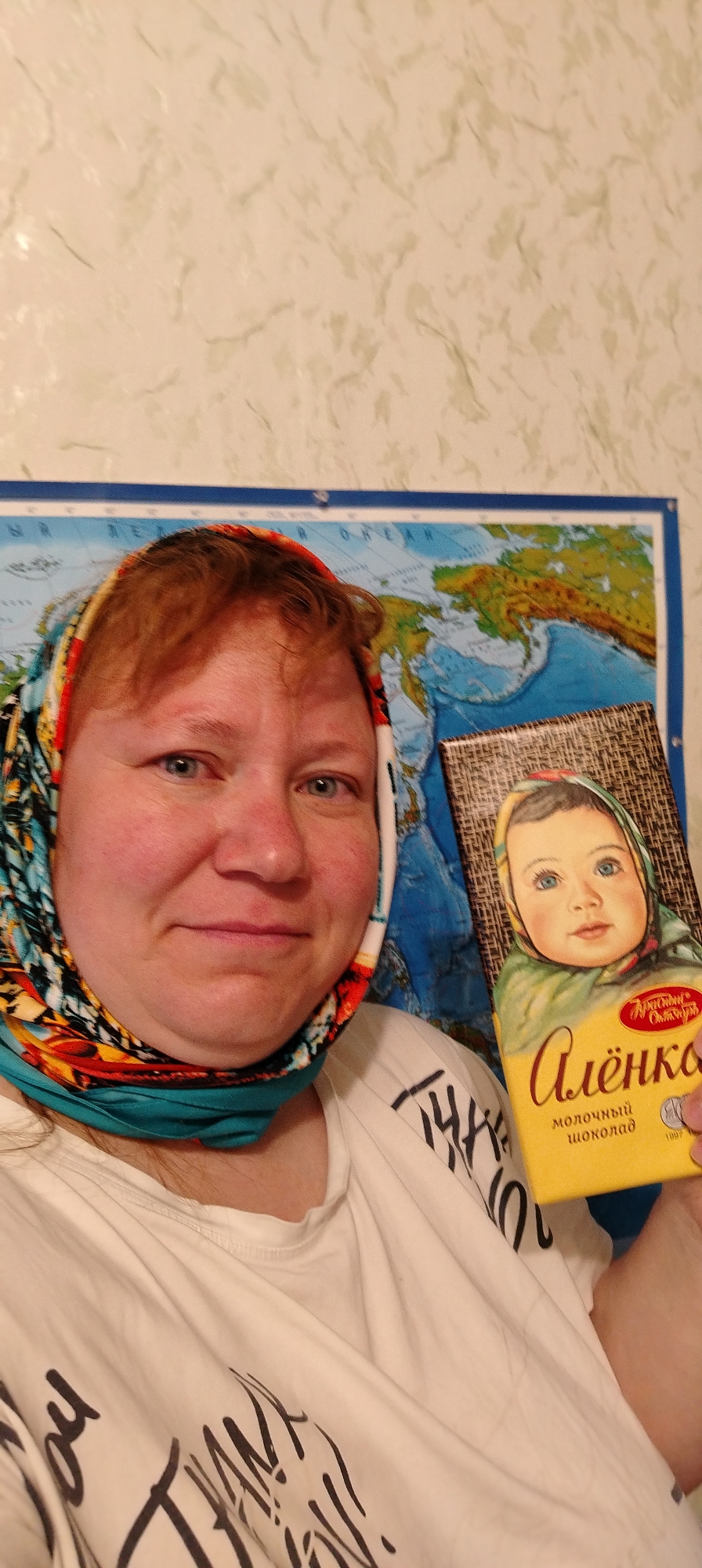 An adult woman, with fair skin, blue eyes, and reddish hair visible at her forehead, is captured in a close-up, chest-up shot. She wears a vibrant headscarf featuring patterns in teal, orange, yellow, and black, wrapped around her head and neck. A white t-shirt with partially visible black stylized text is worn beneath. She holds up an "Alenka" brand milk chocolate bar, its iconic yellow packaging displaying a portrait of a baby in a headscarf. Text visible on the chocolate includes "Алёнка" (Alenka), "молочный шоколад" (milk chocolate), and "Красный Октябрь" (Krasny Oktyabr), with "1967" also present. The woman offers a gentle, pleasant smile directly to the camera. In the background, a world map is affixed to a light-colored wall with a subtle, textured pattern. Portions of landmasses (possibly Europe/Asia) and the Arctic Ocean are discernible on the map, with Cyrillic text "ОКЕАН" (Ocean) visible. The indoor setting, common Russian chocolate, and Cyrillic text strongly indicate a location in Russia, aligning with the provided context of Chaikovsky. The image appears to be a casual selfie, capturing a personal moment.