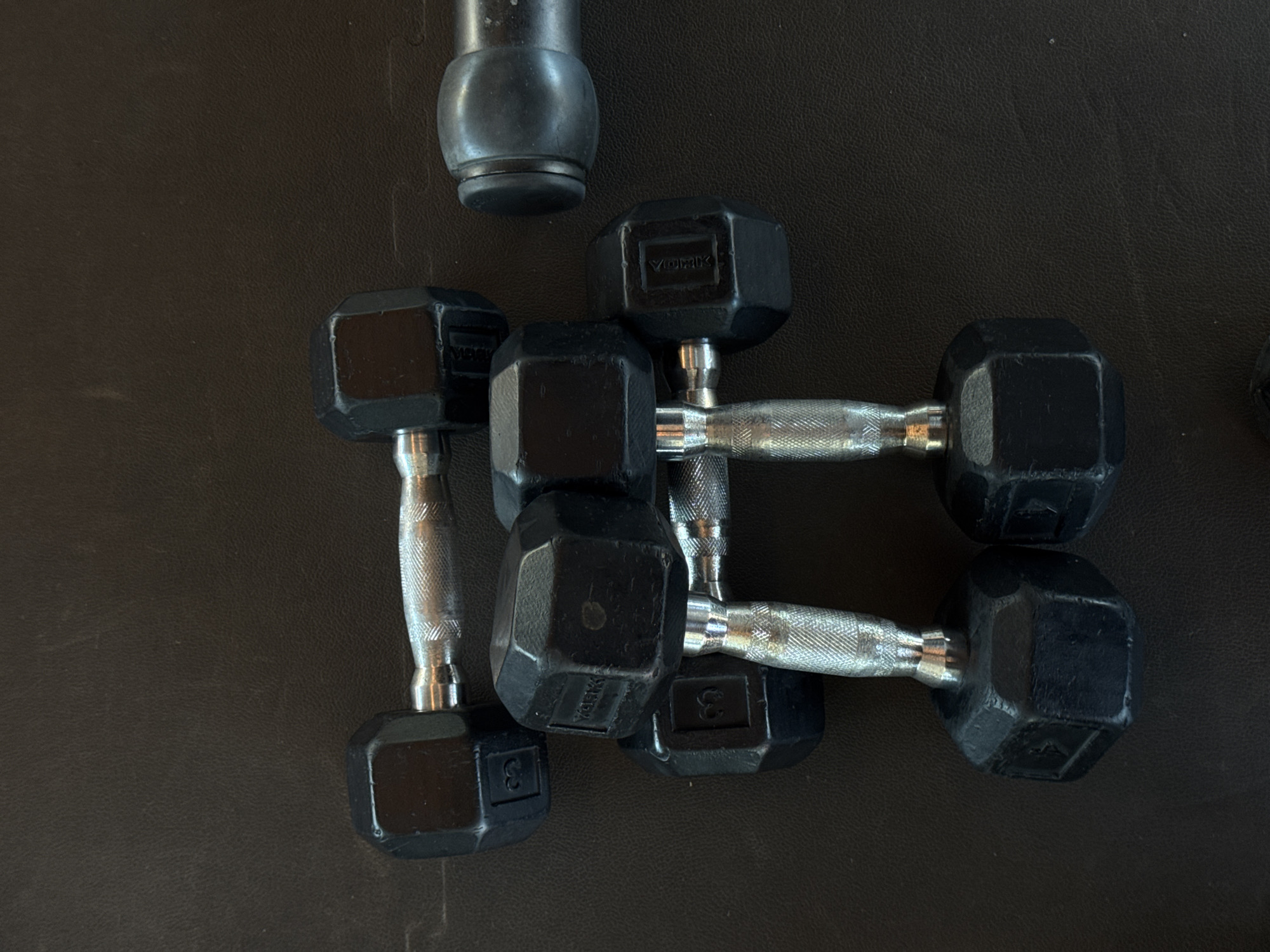 The image shows a set of six black York dumbbells lying on a black, slightly textured surface. The dumbbells are arranged somewhat haphazardly, with some overlapping others. The dumbbells appear to be of varying weights, some with markings indicating "3." There is also an ambiguous small black object with a rounded shape. The environment is a flat, dark surface. The scene appears to be indoors and is lacking any obvious details suggesting Barcelona, Spain.