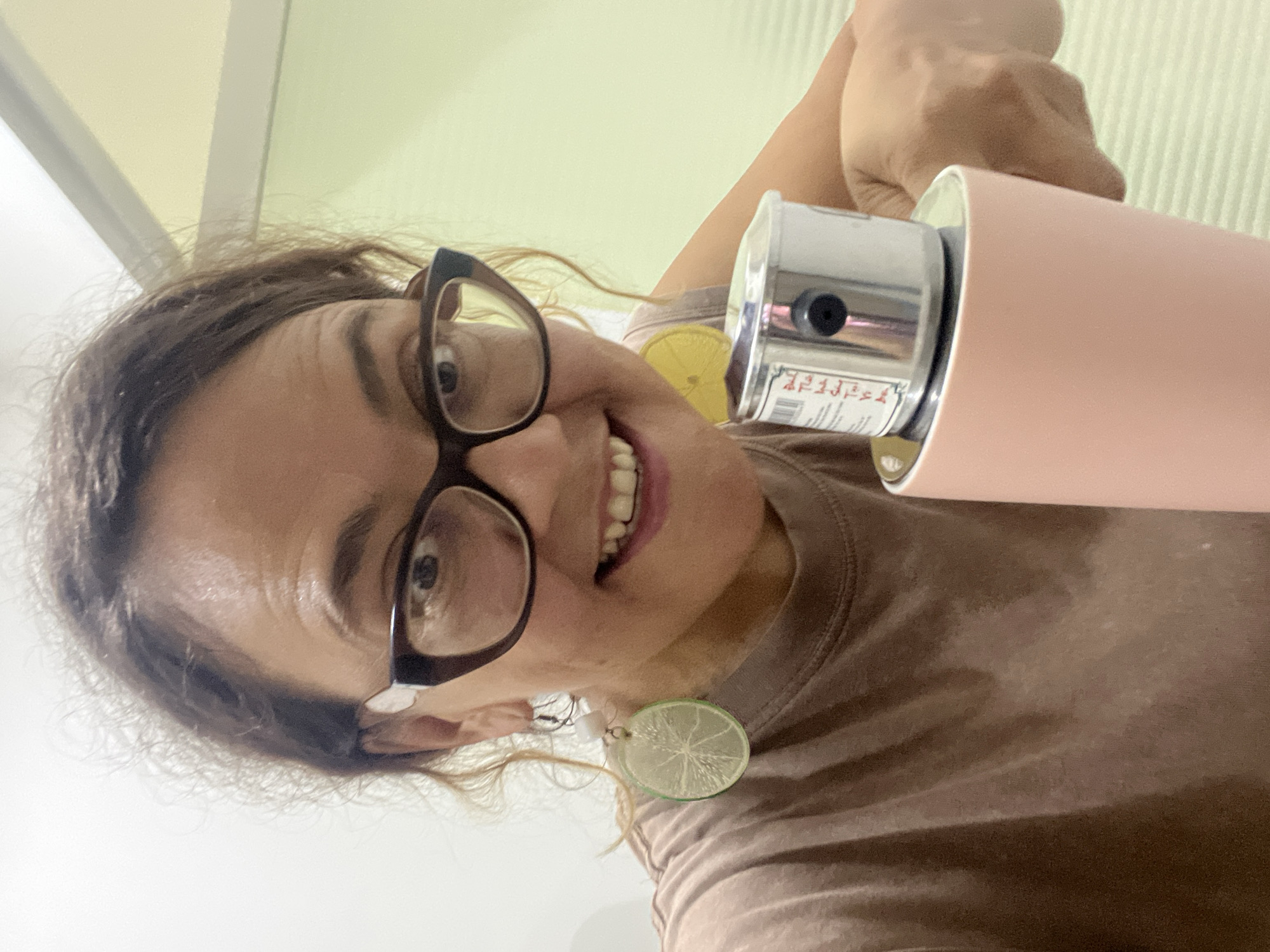 A woman with brown curly hair, wearing glasses and a brown t-shirt, smiles at the camera. She is wearing novelty earrings shaped like lime slices and holding a pink thermos with a silver lid. The background appears to be an indoor setting with a light green wall and a white ceiling. The image is a close-up, likely taken by the woman herself. The location is not specified in the image.