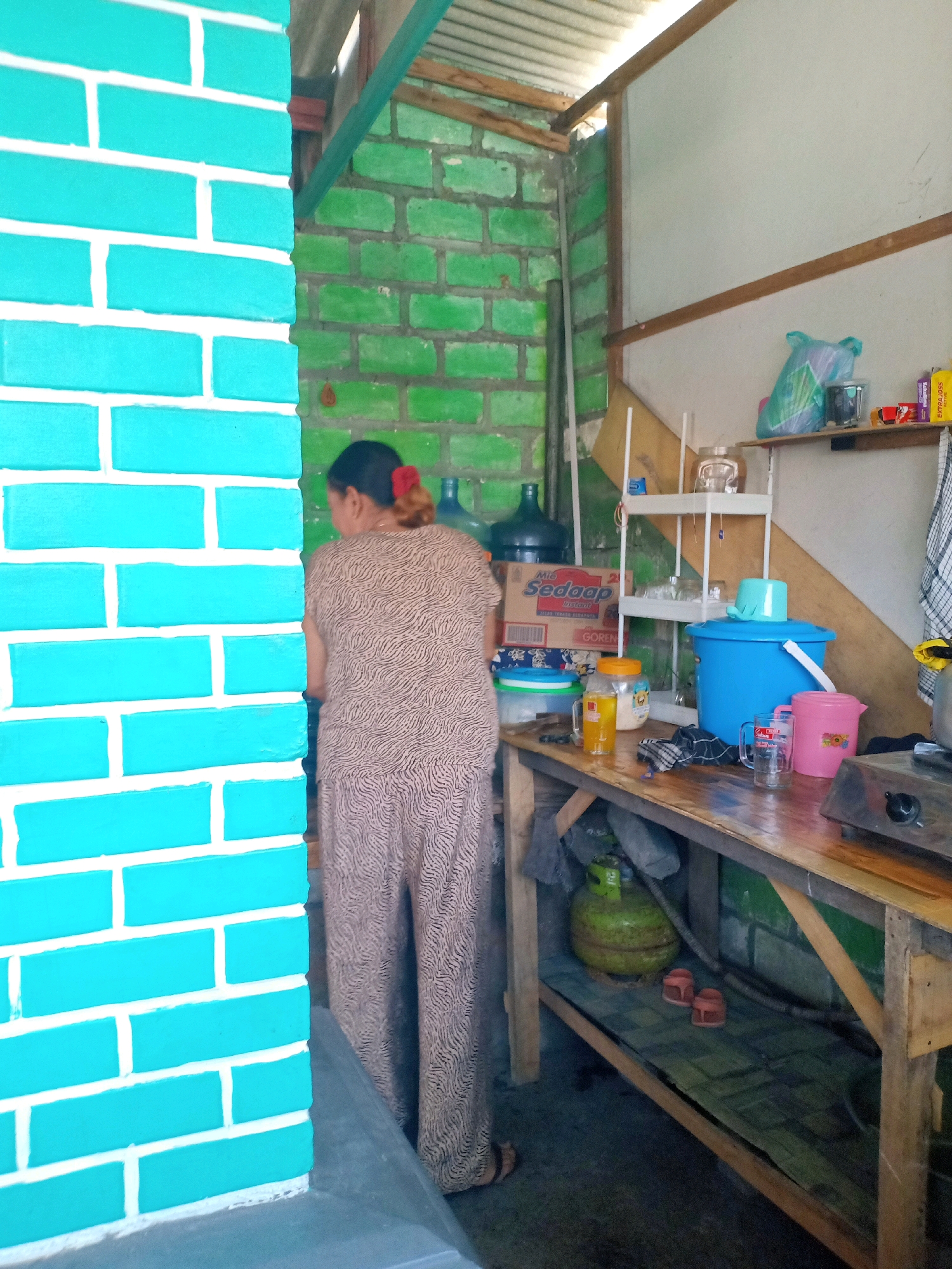 The image depicts an outdoor kitchen area. A woman, viewed from the back, is standing facing a wooden countertop where various items are placed. These include a large blue bucket with a light blue lid, a pink pitcher, a clear drinking glass, and two glass jars, one containing a yellow liquid and the other appearing to hold dry goods. Behind the woman, there are several large water cooler bottles, one dark blue and another clear with water. To the left of the woman is a teal-colored brick wall with white mortar lines. Behind the countertop, there is a shelf holding a blue plastic bag, a smaller clear container, and some boxed items, including a box labeled "Sedaap Mie Goreng". A green gas cylinder is visible on the floor beneath the countertop. The walls of the kitchen area are constructed from rough, green-painted bricks. The ceiling appears to be made of corrugated metal supported by wooden beams. The scene is likely situated in Sumurgung, Indonesia, based on the context provided.