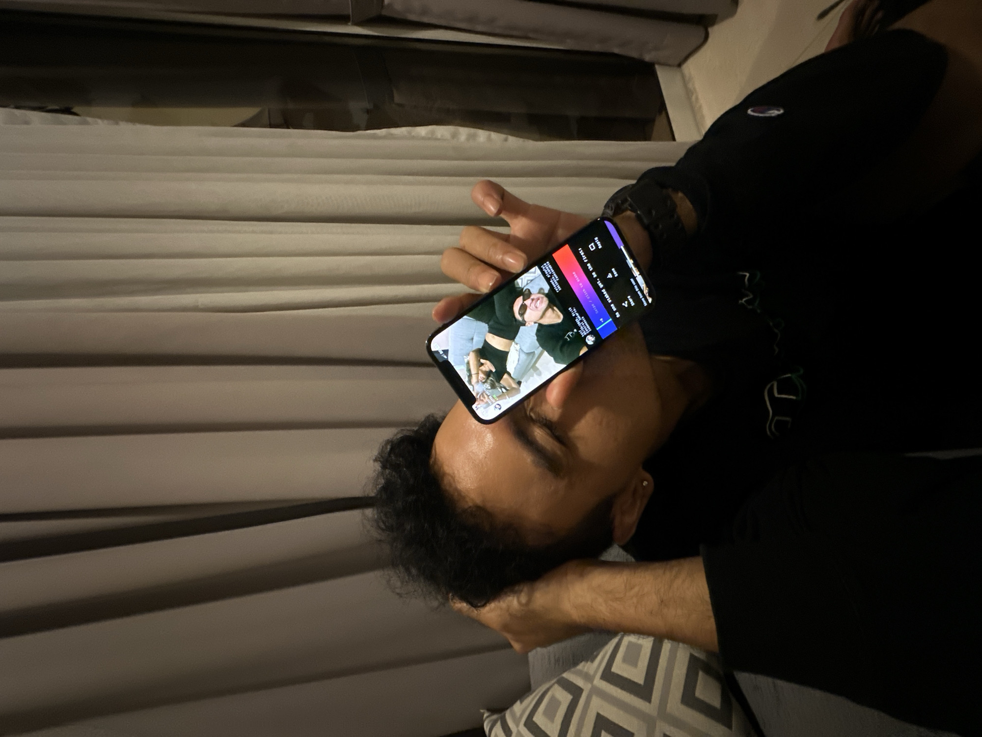 The image shows a man lying down, likely on a bed or sofa, with his head resting on his hand. He is holding a smartphone directly above his face, displaying a video or image. The phone screen shows a person with sunglasses. The man is wearing a black shirt with a visible logo on the upper chest. He also has a watch on his wrist. The background includes a window with blinds or curtains. A patterned pillow can be seen near his arm. The location is not discernible from the image and cannot be placed in Central Jakarta, Indonesia.