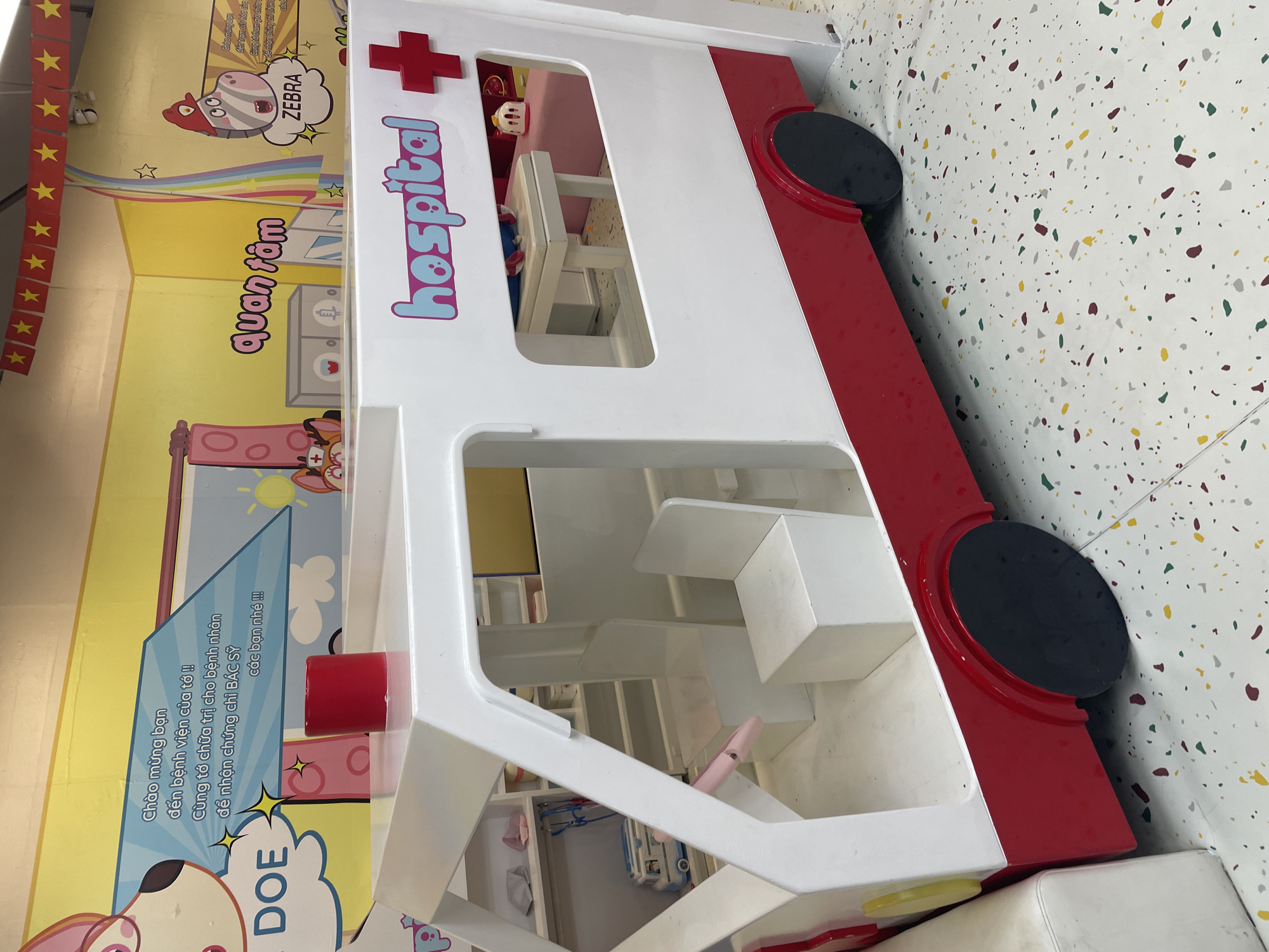 The image depicts a child's play structure resembling an ambulance or hospital setup. It is painted in white and red with features like wheels, a "hospital" sign, and a plus sign symbol typically associated with healthcare. The interior of the structure is visible, revealing shelves and play furniture. The background includes a decorated wall with cartoon-like illustrations of animals and text in Vietnamese. The text includes phrases such as "chào mừng bạn" which means "welcome" and refers to a "zebra." The setting is likely within a child care facility or entertainment center in Nha Trang Ward, Vietnam. The speckled flooring is also visible.