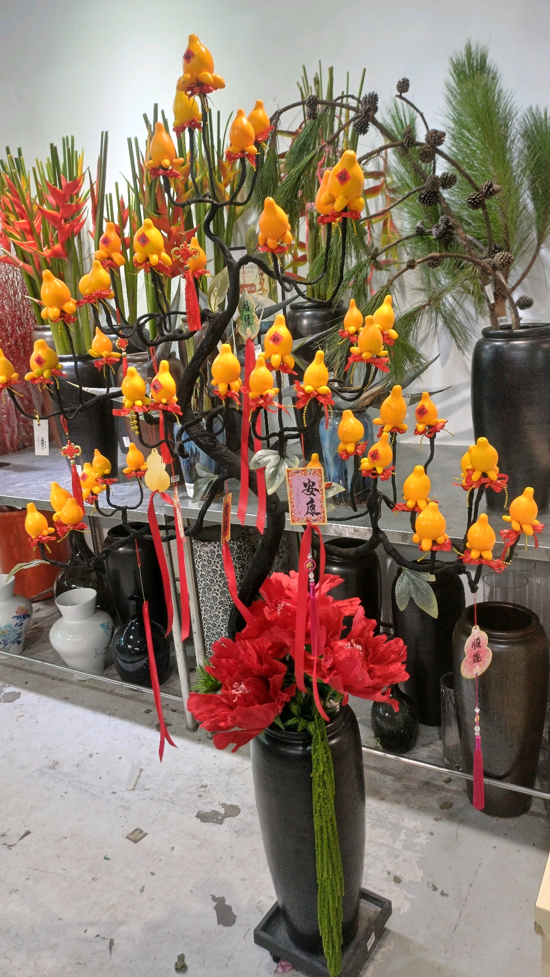 The image depicts a floral arrangement featuring artificial yellow gourds with red tassels, resembling a decorative tree, on display in what appears to be a shop or market stall. Behind the main arrangement, there are various potted plants, including some with red heliconia flowers and others with pine needles and cones. Several dark-colored vases are visible in the background and foreground. In the foreground, a large black vase holds a bouquet of vibrant red flowers, with a green decorative element hanging down its side. Decorative Chinese paper tags with red Chinese characters are attached to some of the branches and vases, suggesting a festive or celebratory theme, likely related to Chinese New Year. The overall scene is well-lit, with a white background that might be a wall. The flooring is a light-colored, textured surface, possibly concrete, with some scattered debris. There are no people visible in the image. Without additional context or specific visual cues that definitively place it within Kuala Lumpur, Malaysia, such as street signs, distinct architectural elements, or recognizable landmarks, the precise location cannot be determined. However, the style of the decorations and the presence of Chinese characters strongly suggest a setting within a community that celebrates Chinese culture, which is prevalent in Malaysia, including Kuala Lumpur.