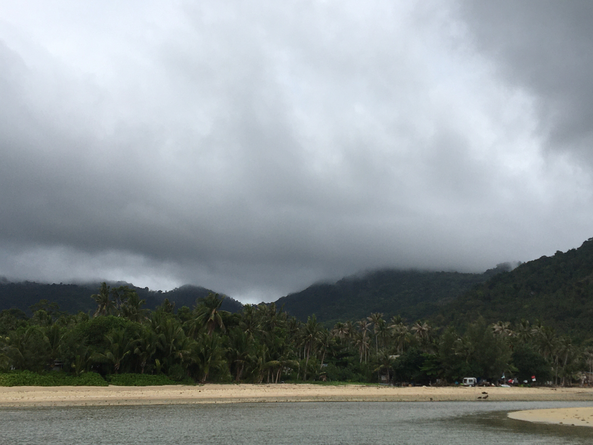 The image presents a coastal landscape under a heavy, overcast sky. The scene is likely in Baan Kai, Thailand, given the tropical vegetation and geographical features.
In the foreground, there is a body of water, likely a lagoon or bay, leading to a sandy beach. The beach is relatively wide and appears to be lightly populated, with a few indistinct figures in the distance.
Behind the beach, dense tropical vegetation is present, comprised of palm trees and other greenery. This vegetation stretches inland towards the base of hills or low mountains. The hills are covered in similar dense vegetation, and their peaks are partially obscured by low-hanging clouds.
A few structures and a white vehicle, possibly a camper van, are visible among the trees on the right side of the image.