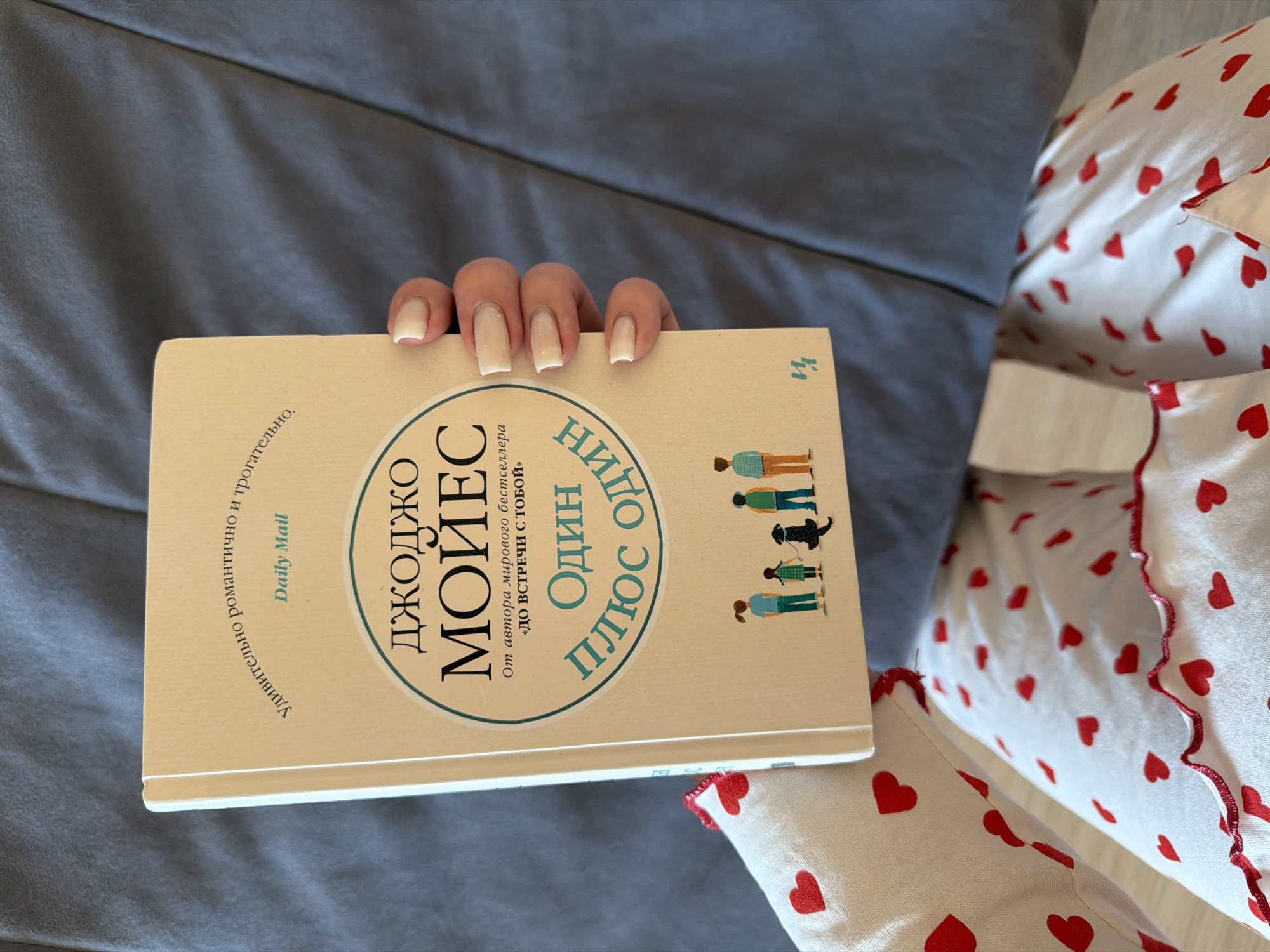 The image shows a hand holding a book on a bed or couch. The book cover has text in Russian, including the author's name "Джоджо Мойес." The text also mentions "Daily Mail." The bedclothes have a pattern of red hearts on a white background. The location is not specified, and Chaikovsky, Russia cannot be confirmed from the image.