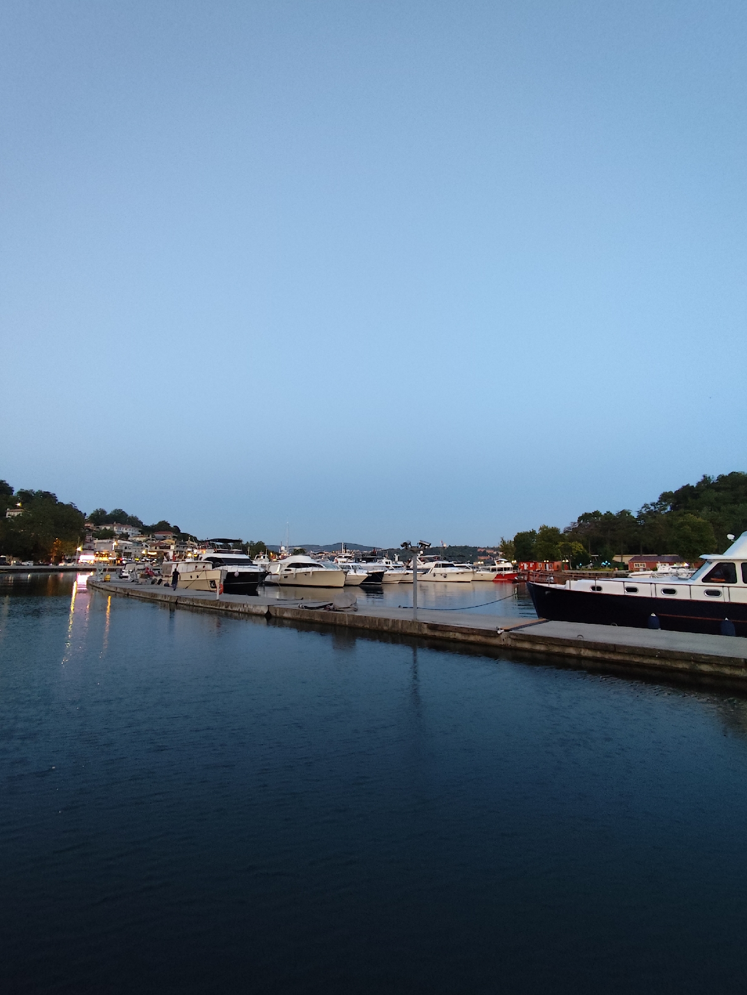 The image depicts a tranquil harbor scene at dusk, likely in Sarıyer, Turkey, given the presence of boats and a hillside populated with buildings. The sky is a soft, clear blue, transitioning into twilight. In the foreground, dark, calm water reflects the faint lights from the shore. A concrete pier extends into the water, lined with several white and dark-hulled boats of various sizes, including what appears to be a sleek motor yacht on the right. A few individuals are visible on the pier, engaged in relaxed activity. The background features a lush green hillside dotted with houses and buildings, with some lights illuminated, suggesting the presence of a town or village. The overall atmosphere is serene and peaceful, characteristic of a harbor at the end of the day.