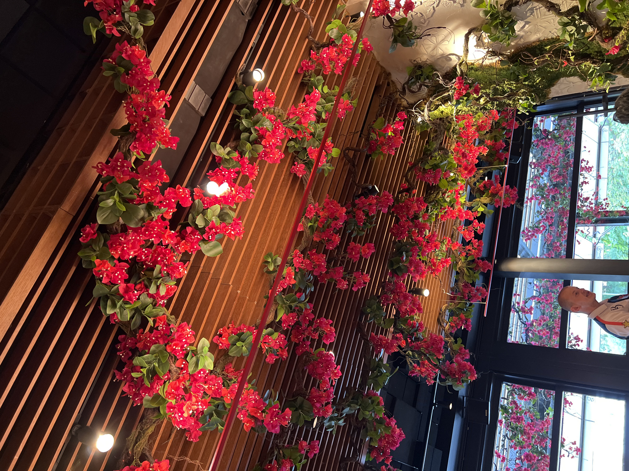 The image shows a restaurant ceiling adorned with artificial red bougainvillea flowers and green foliage. The ceiling is made of wooden slats, and spotlights are intermittently placed. To the left, there are large windows revealing a glimpse of the exterior with more flowers. In the foreground, a man with short hair and a collared shirt with colorful trim is visible from the chest up, facing right and speaking. The overall impression is a vibrant and decorated interior space. Based on external information, this scene is from the restaurant "Boom! Burgers" located in Sofia, Bulgaria.