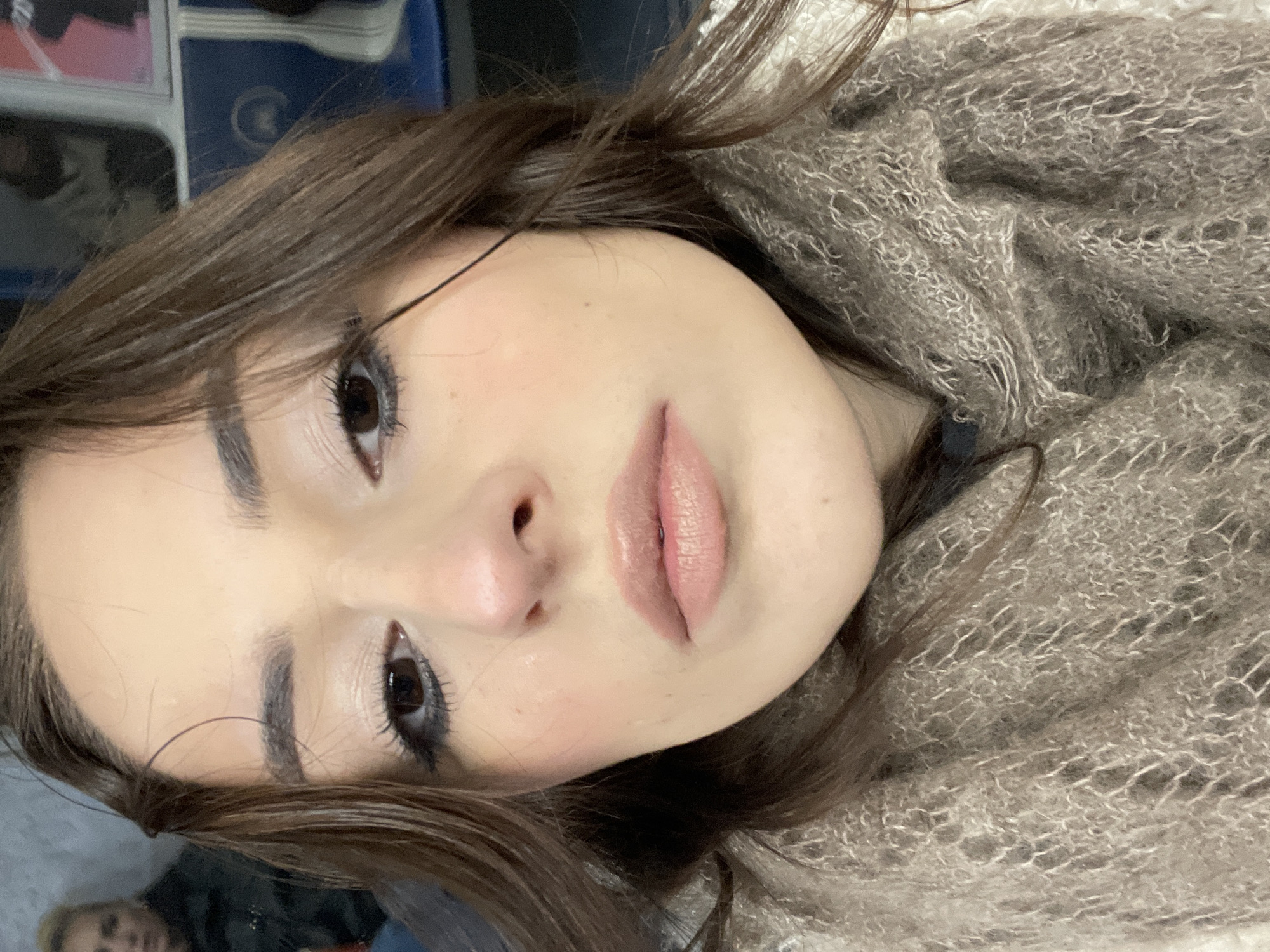 A close-up portrait features a young woman, likely in her late teens or early twenties, looking directly into the camera. She has long, dark brown hair that cascades around her face and over her shoulders. Her dark eyes are accentuated with eyeliner and mascara, complementing her well-groomed, dark eyebrows. She wears a light pink or nude lipstick and subtle blush on her fair skin.
The woman is wrapped in a voluminous, light brown or taupe shawl or scarf, characterized by a fuzzy, loosely woven texture. The fabric covers her neck and parts of her chest and shoulders, suggesting a warm garment. The image is framed at a slight angle, making it appear as if she is reclining or leaning back. Her expression is neutral and calm.
The background is softly blurred but suggests an indoor setting, with indistinct shapes of blue and white objects, possibly furniture or storage. A faint, blurred reflection of another individual with blonde hair can be glimpsed in the lower left corner, indicating the presence of a reflective surface such as a mirror. The lighting is soft and even, highlighting her facial features. There is no visible text in the image. The location context for this scene is Moscow, Russia.