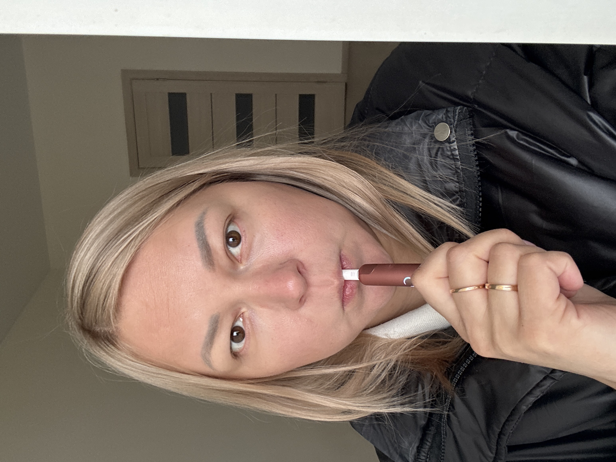 A fair-skinned woman with blonde hair is shown from the chest up, holding a brown electronic device in her mouth. She wears a black puffer jacket and has two gold rings on her fingers. Behind her is a white wall and a door with three vertical panels. There is no indication of location in the city of Izhevsk, Russia.