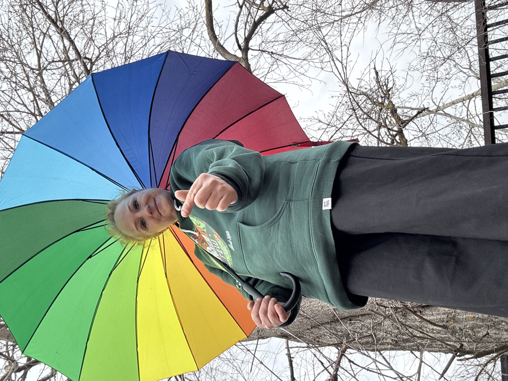 A woman stands beneath a rainbow umbrella in Chaikovsky, Russia, against a backdrop of bare trees and an overcast sky. She is wearing a green hooded sweatshirt and black pants, and her face is visible as she looks down at the camera. A portion of a ladder can be seen in the background amidst the branches.