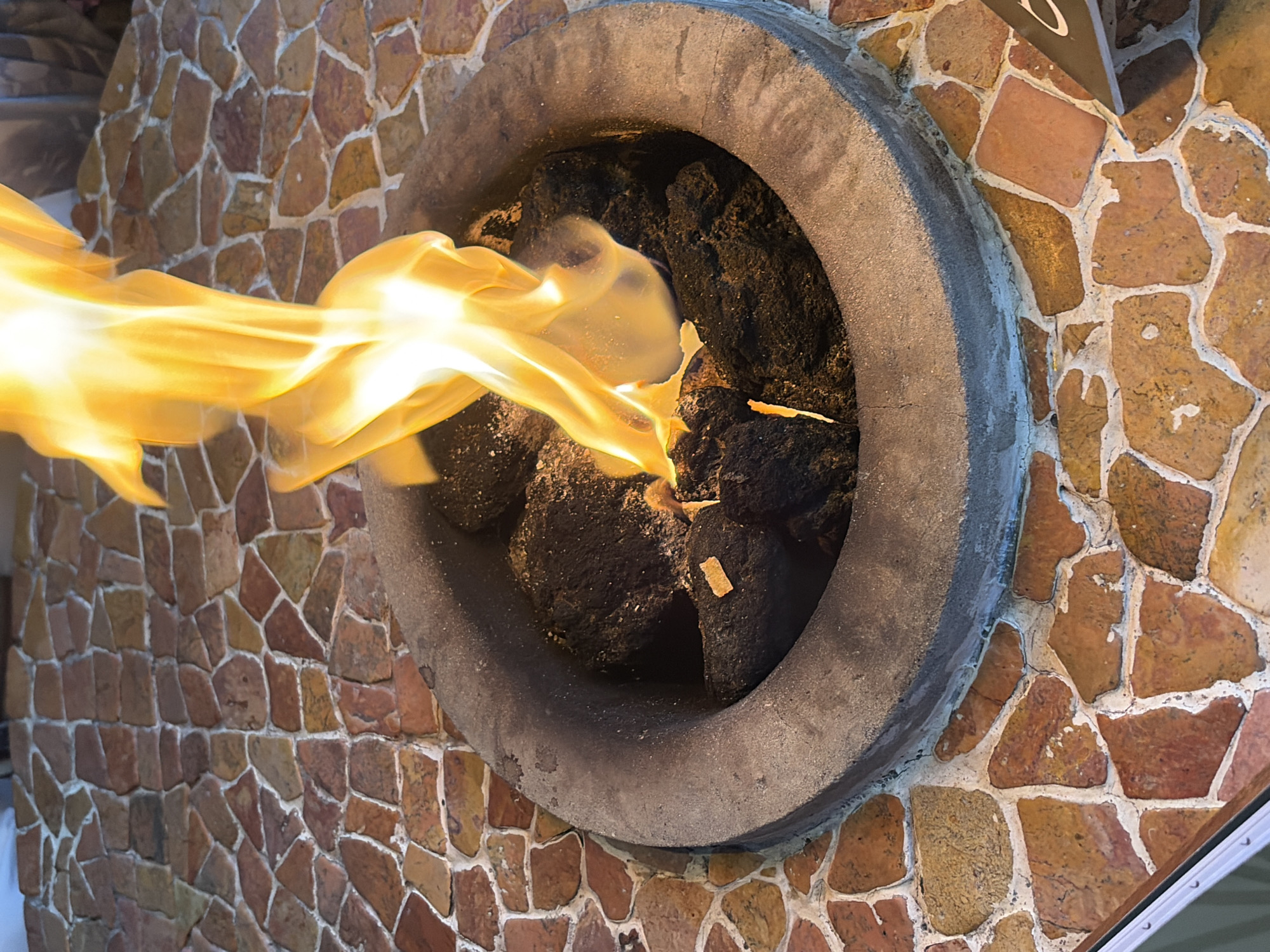 An outdoor fire pit is centrally located in the frame. The fire pit is a circular, dark gray, concrete basin filled with dark, volcanic rocks. Bright orange and yellow flames leap from the center of the rocks. The fire pit is surrounded by a mosaic tabletop made of irregularly shaped, multi-colored stones, predominantly shades of reddish-brown and tan, with white grout. The tabletop is set in an outdoor environment, likely a patio or deck, as suggested by the patterned stone surface. The image appears to be taken in Kintamani, Indonesia, a region known for its volcanic landscapes and outdoor dining experiences. No people are visible in the image.