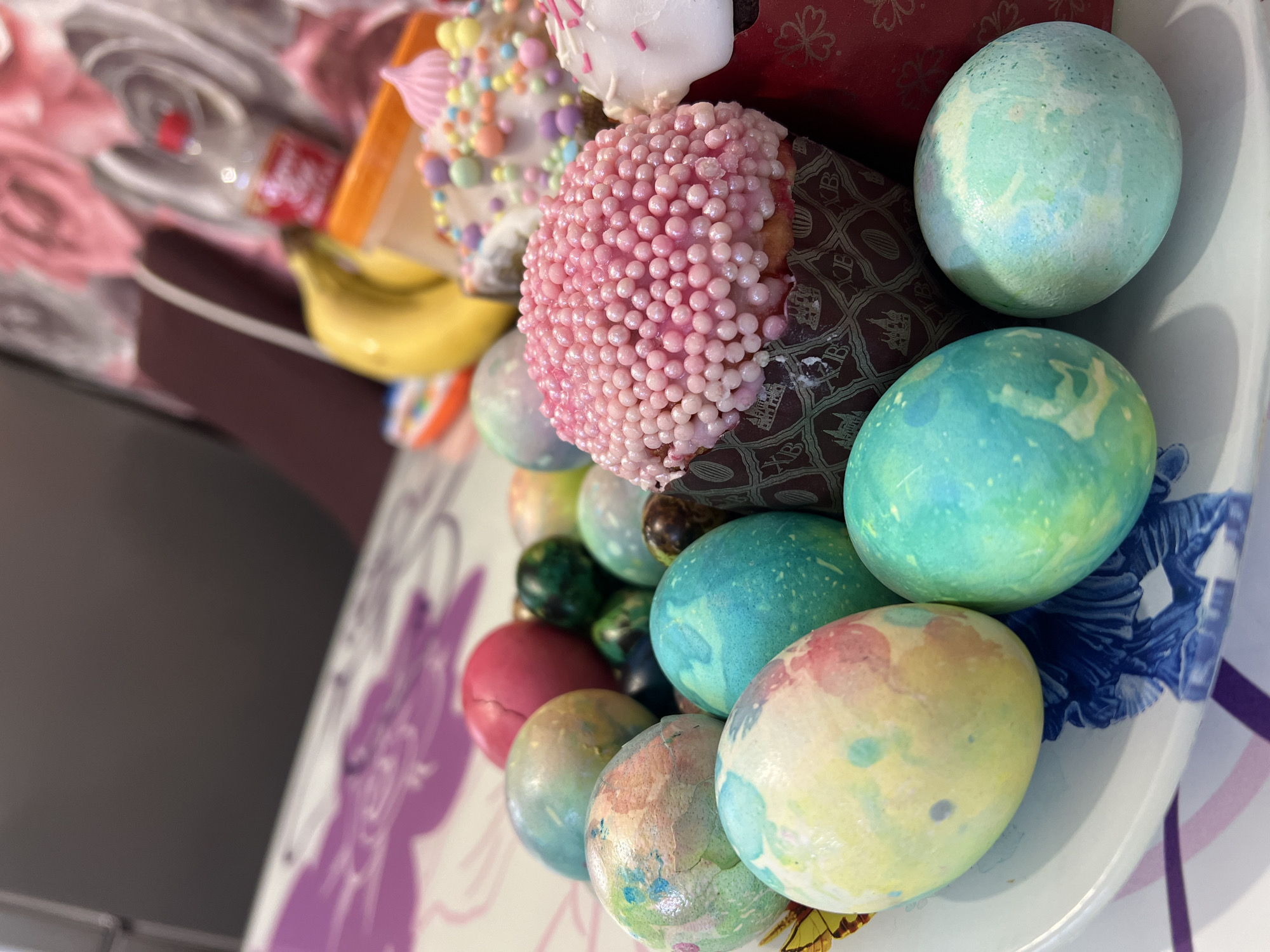 A high-angle shot displays a plate of decorated Easter eggs and Kulich cakes. The eggs are dyed in various colors, including blue, green, yellow, and red, with a mottled or marbled effect. The Kulich cakes are decorated with white, pink, and multi-colored sprinkles or icing. Some of the cakes are in patterned wrappers. A banana is visible among the cakes. The plate has a blue floral pattern on the edge and a white rim. The background features a blurry purple and white surface, potentially a tablecloth. The scene likely takes place indoors. The location in the city of Noviy, Russia is not discernible from the image.