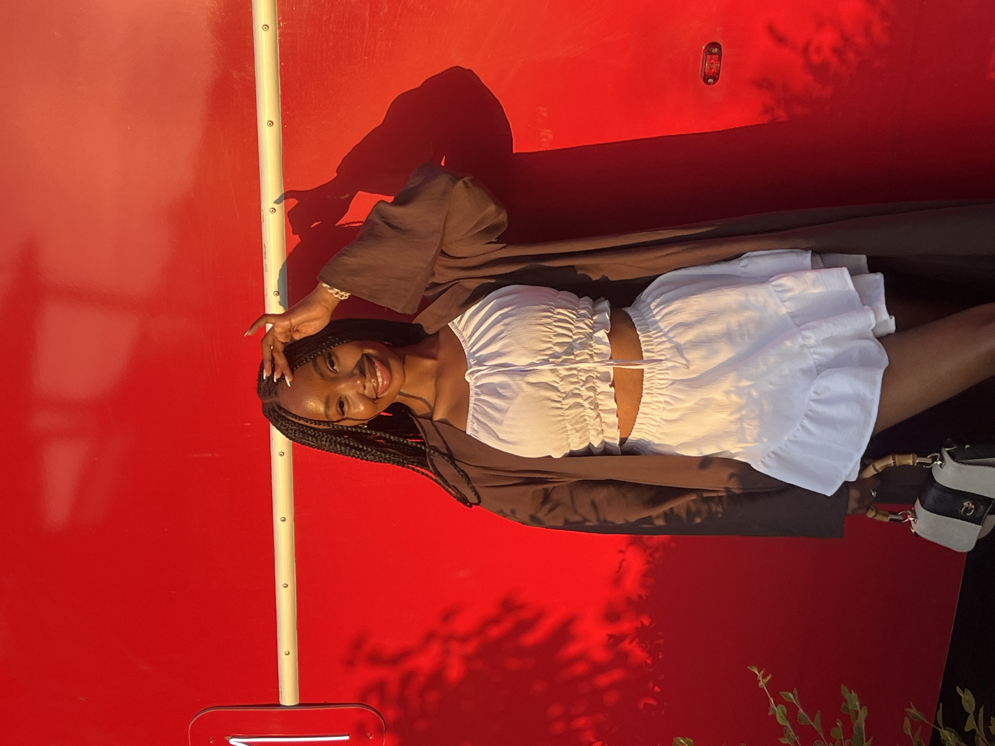 A young Black woman with braided hair smiles at the camera, holding her right hand to her forehead. She is wearing a white ruffled crop top and a matching white ruffled skirt, layered under a dark brown open cardigan. She carries a black and white handbag with bamboo handles. The woman is standing against a bright red wall, possibly part of a building or vehicle, which has a horizontal yellow stripe running across it. Shadows of leaves and branches are visible on the red surface. A small red light fixture is present on the right side of the wall. The scene appears to be outdoors during daylight, with warm lighting casting shadows. The location is identified as Kazan, Russia.