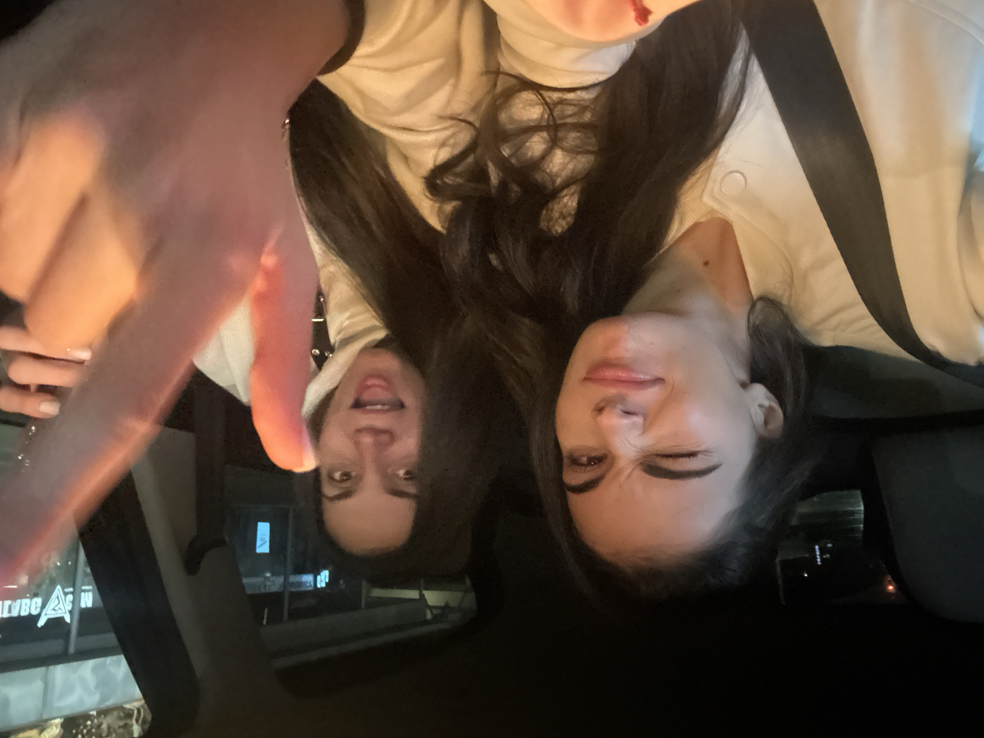 In an indoor environment, two young women are seated inside a vehicle. The woman on the left is winking, her left eye closed and her right eye open. She is looking towards the camera, with a slight smile on her face. The woman on the right has her mouth open in a surprised expression, looking towards the camera as well. Her right hand is raised, with her thumb and index finger extended, pointing towards the camera. The background shows a nighttime cityscape with illuminated buildings. The woman on the left is wearing a white jacket with a black seatbelt across her chest. The woman on the right is wearing a light-colored hooded sweatshirt. The car interior is dark, with hints of interior lighting and reflections from the outside. The overall scene captures a candid moment of the two women interacting within the vehicle.