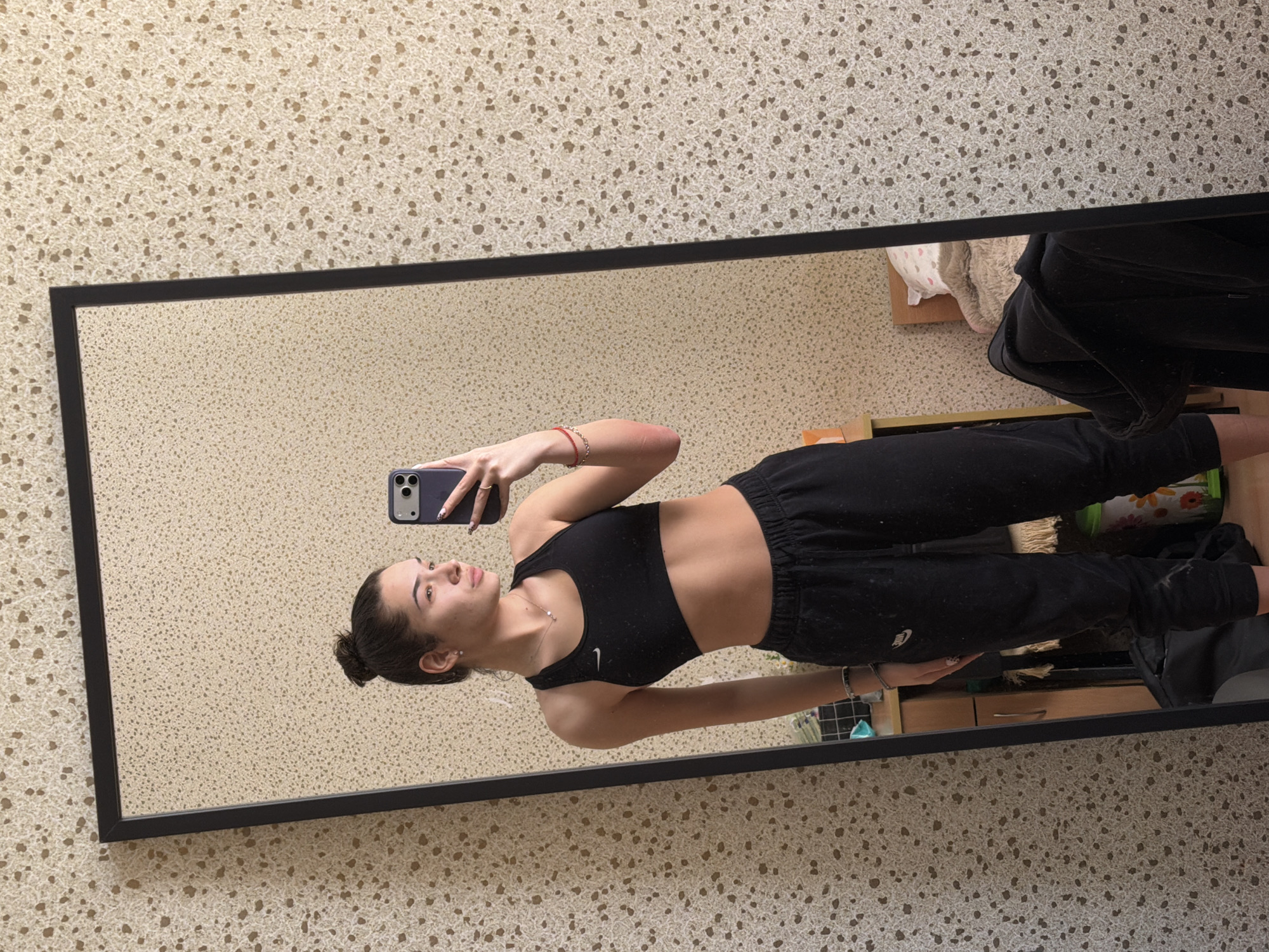 A young woman is reflected in a full-length mirror. She is fair-skinned with dark hair pulled into a bun. She is wearing a black sports bra and black sweatpants. She is holding a smartphone, presumably taking the picture. The reflection shows a room with speckled, beige-toned wallpaper. Other visible items include clothing draped over furniture and a dresser. There is no information to determine the city of Sofia, Bulgaria, as the environment does not provide any specific markers.