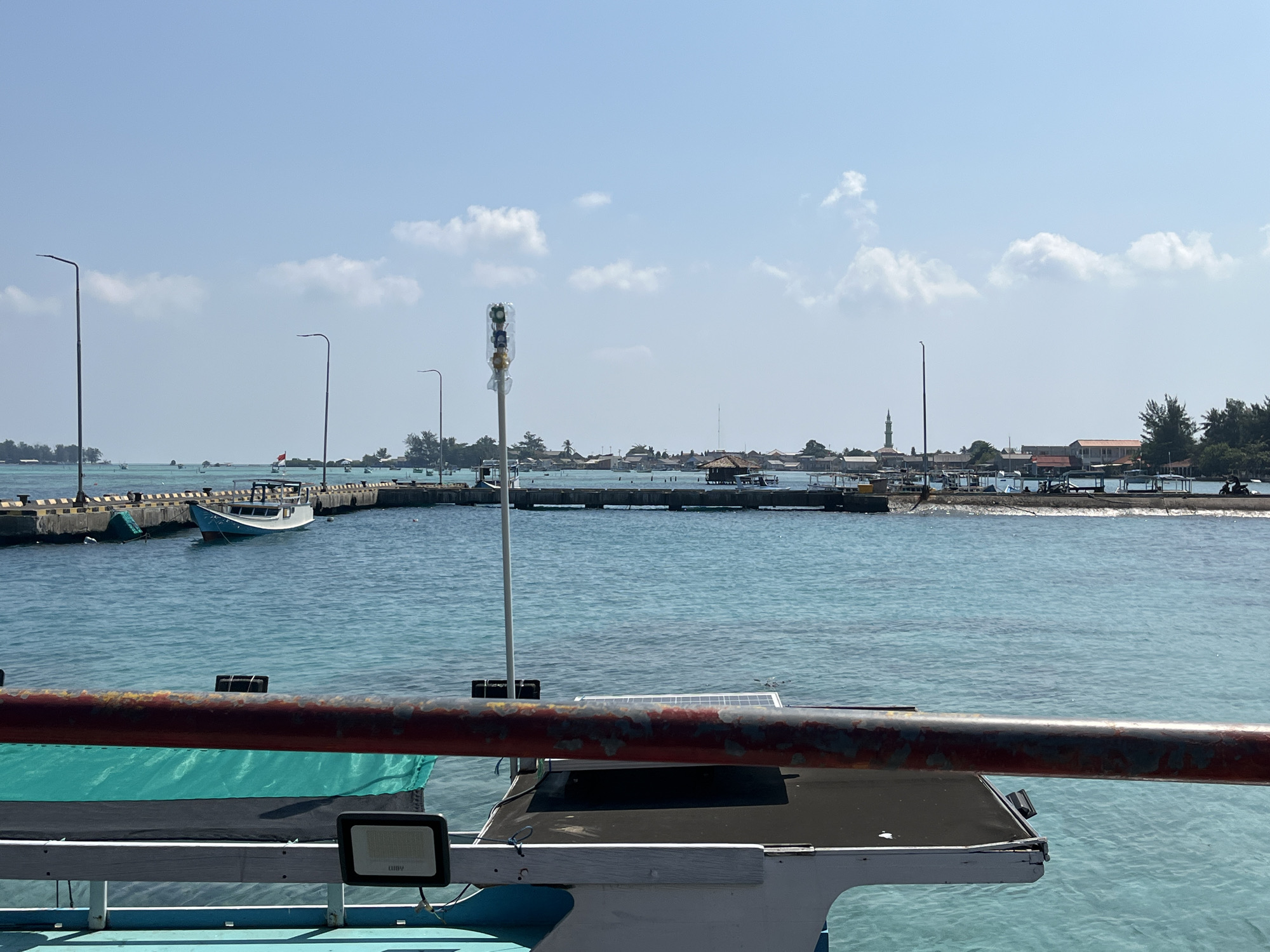 The image shows a bright, sunny day at a harbor in Karimunjawa, Indonesia. The foreground features a part of a boat with a rusty red railing and a green tarp. The water is a clear, turquoise blue. In the mid-ground, a pier extends into the water with several boats docked along it, including a white and blue boat. Streetlights line the pier. The background shows the coastline of Karimunjawa with buildings, trees, and a mosque with a tall minaret. Several other boats are visible in the distance. The sky is clear with a few scattered white clouds.