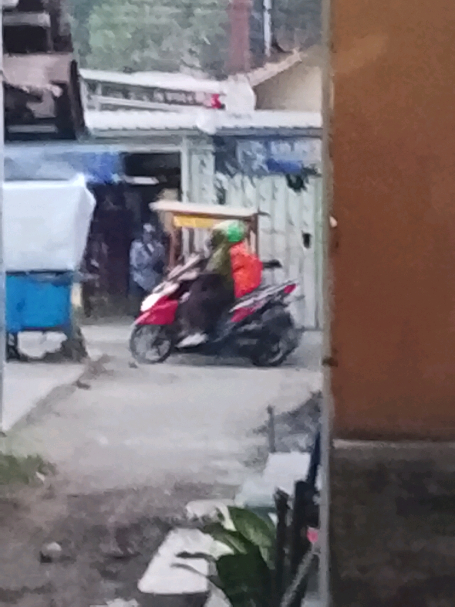 A person, wearing a green headscarf and an orange garment, is riding a red scooter. The scooter has a dark-colored seat and is moving forward. The scooter has a visible front wheel and a rear wheel. A person in a light-colored shirt is visible behind the scooter, standing next to a blue object. The background features buildings with corrugated metal roofs and some greenery. The scene appears to be on a street or alleyway. The location is Ujungrusi, Indonesia.