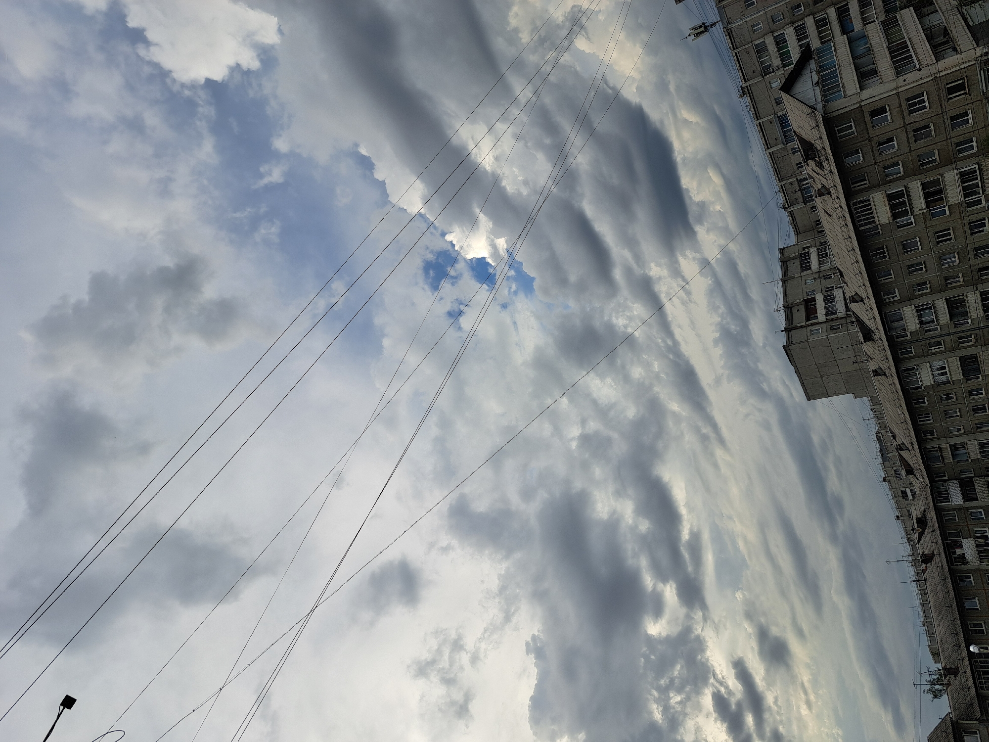 The image captures a slightly overcast sky above a dense urban landscape, likely in Irkutsk, Russia, given the architectural style of the buildings. Multiple sets of overhead wires crisscross the sky, some taut and others with a slight sag. The clouds are a mix of dark gray and lighter, wispy formations, with patches of blue sky visible, particularly in the upper right quadrant.
Below the sky, a series of apartment buildings, characteristic of Soviet-era architecture, dominate the scene. These buildings are grey and utilitarian, with numerous windows and balconies. The roofs are covered in dark, segmented material, and some have visible vent pipes or antennae. The perspective suggests the photo was taken from a higher vantage point, looking down onto the upper floors and rooftops of the buildings. There are no people or vehicles visible in the frame. The overall impression is one of a typical urban day in a residential district.