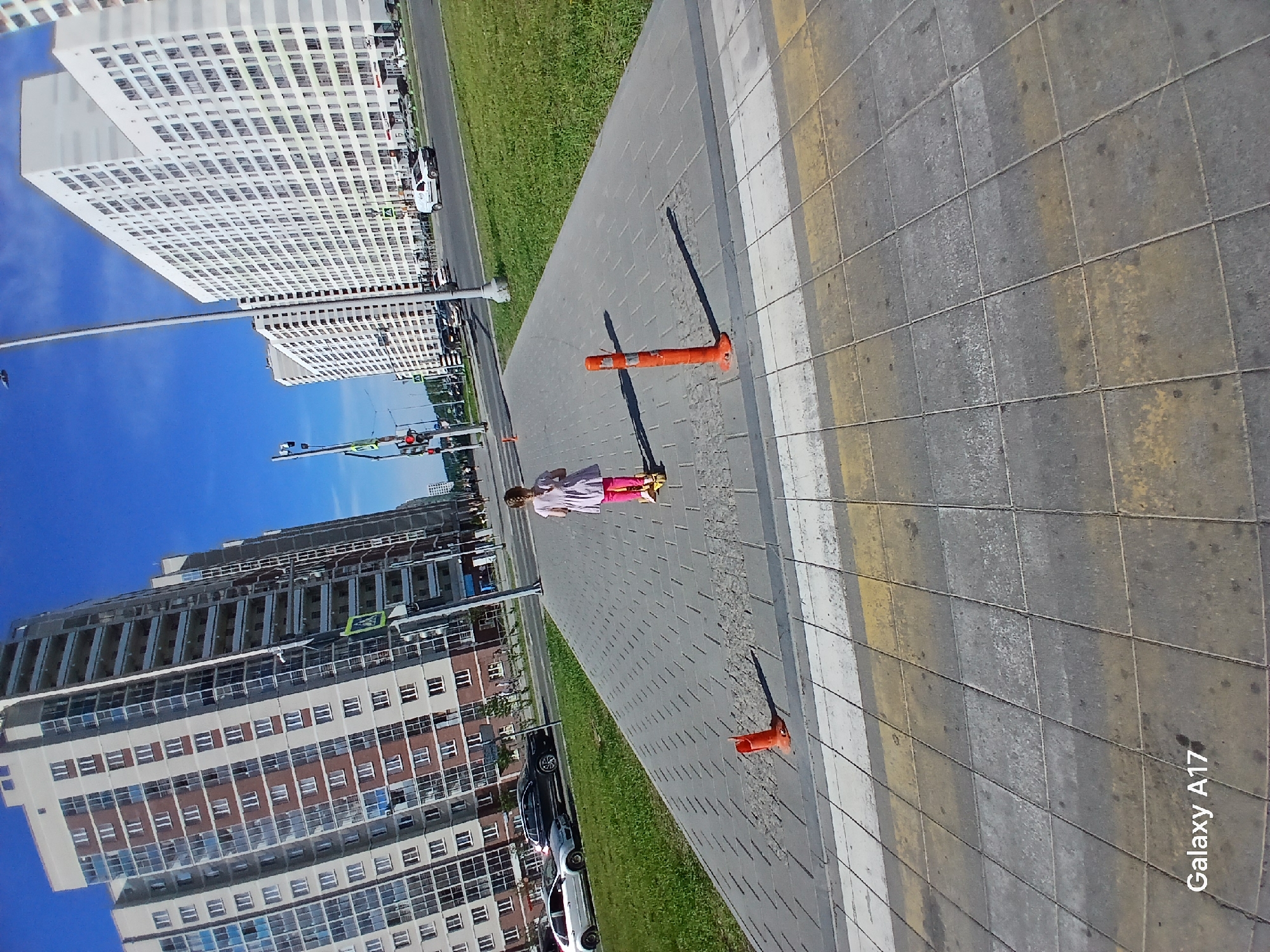 The image shows an outdoor scene on a sunny day. A girl is walking on a paved sidewalk lined with grass. On one side of the sidewalk, there are several high-rise buildings with multiple windows. On the other side, there's a road with parked cars. There are also two orange traffic cones on the sidewalk. Based on the buildings and the urban layout, the location may be in Yekaterinburg, Russia.