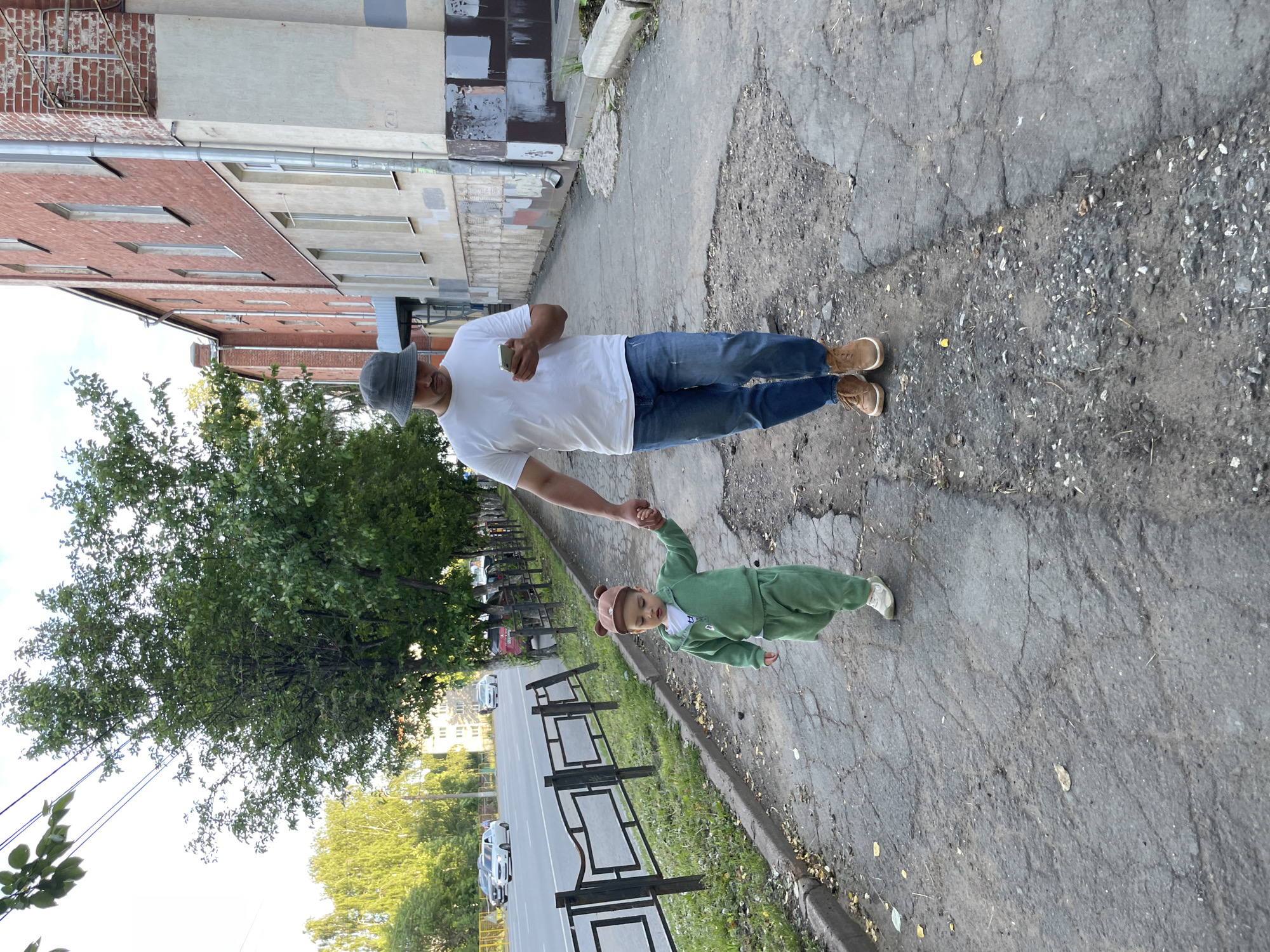 The image depicts a daytime scene in Izhevsk, Russia, featuring an adult man and a young child walking hand-in-hand on a cracked asphalt sidewalk. The man, positioned on the left, wears a white short-sleeved t-shirt, blue jeans, and tan boots, topped with a gray bucket hat. He holds a smartphone in his left hand while extending his right hand to grasp the child's hand.
The child, a toddler or young child, is dressed in a matching green full-body outfit and a brown hat with small, ear-like details. They wear light-colored sneakers, and their eyes appear to be closed or looking downwards, suggesting they might be sleepy or looking at the ground.
The sidewalk they are traversing is uneven and severely cracked in places, with gravel and small debris visible. To the right of the sidewalk, a narrow strip of grass is bordered by a decorative black metal fence. Beyond the fence lies a road with several parked or moving cars, mostly light-colored sedans. A dense line of green trees runs along the opposite side of the road, and more trees are visible in the distance.
On the left side of the frame, a tall, multi-story building constructed of red bricks dominates the background, featuring numerous windows and a visible downspout. Further back and to the right, another building with a lighter, possibly beige or grey, facade and distinctive horizontal rectangular window structures can be seen. The sky above appears bright, indicating clear weather conditions during the day. No discernible text is visible on any objects or surfaces. The overall setting suggests an older urban or suburban environment.