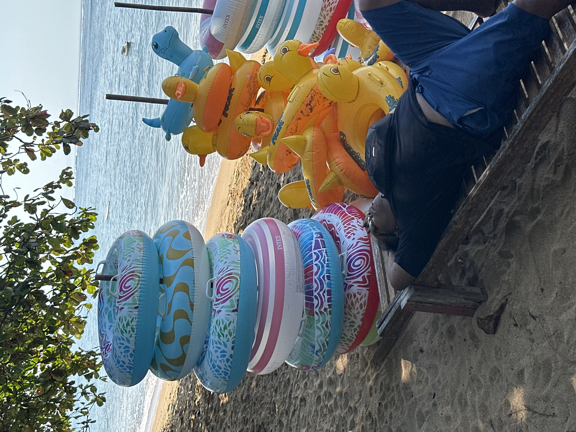 In Denpasar, Indonesia, on a sandy beach, a person reclines on a wooden lounge chair under the shade of a leafy tree. They are wearing a black t-shirt and blue shorts. In front of the lounge chair, a stack of inflatable swimming rings with various colorful patterns is arranged, and to the right, a collection of inflatable animal-shaped floats, including yellow ducks and a blue dinosaur, are piled. The ocean is visible in the background with gentle waves lapping at the shore.