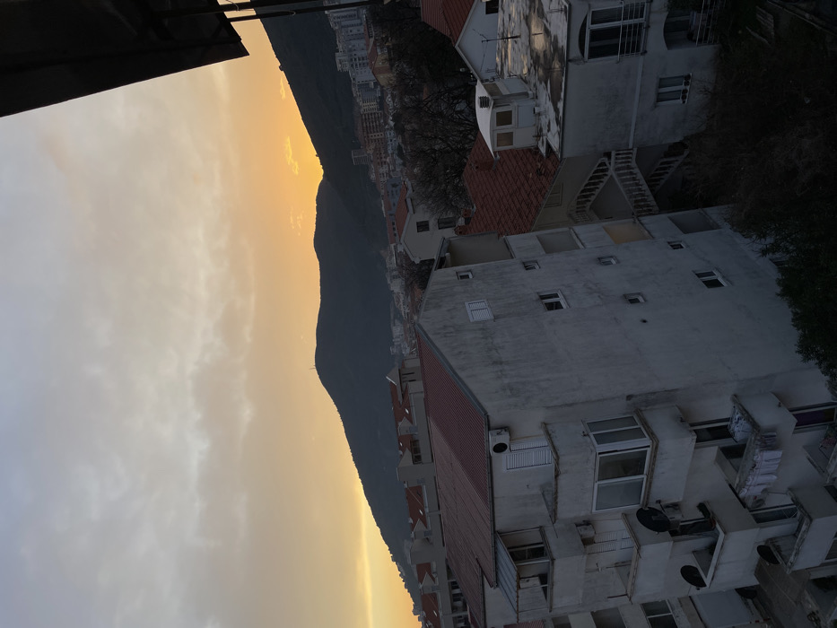 The image depicts an urban landscape in Budva, Montenegro, viewed from an elevated vantage point during sunset or dusk.
The sky occupies the upper portion, exhibiting a gradient from light grey-blue at the zenith to a vibrant orange-yellow at the horizon, indicative of the setting sun. Scattered, thin clouds are present across the sky.
A dark, prominent mountain range forms the distant background, silhouetted against the bright horizon. A slender, dark pole-like structure, possibly an antenna or communication tower, is visible near the summit of the central mountain.
The midground and foreground are dominated by a dense arrangement of multi-story residential buildings, characterized by light-colored (off-white or grey) facades and uniformly red-tiled gabled roofs.
In the left foreground, a five-story apartment building features multiple balconies with white railings. Two black satellite dishes are mounted on the exterior. Several external air conditioning units are affixed to the facade. On a lower balcony, white laundry items are visibly hanging on a clothesline. The building also displays various windows, some with closed white louvered shutters.
To the right of this building, another structure with a similar facade and a red-tiled roof is visible. An external, white-railed staircase descends between this building and an adjacent lower structure, which features a flat, wet-appearing roof and a distinct, small white-enclosed extension on its rooftop.
Sparse, deciduous trees with bare branches are interspersed among the buildings, and some evergreen foliage is visible at the bottom of the frame. No individuals are visible in the scene.