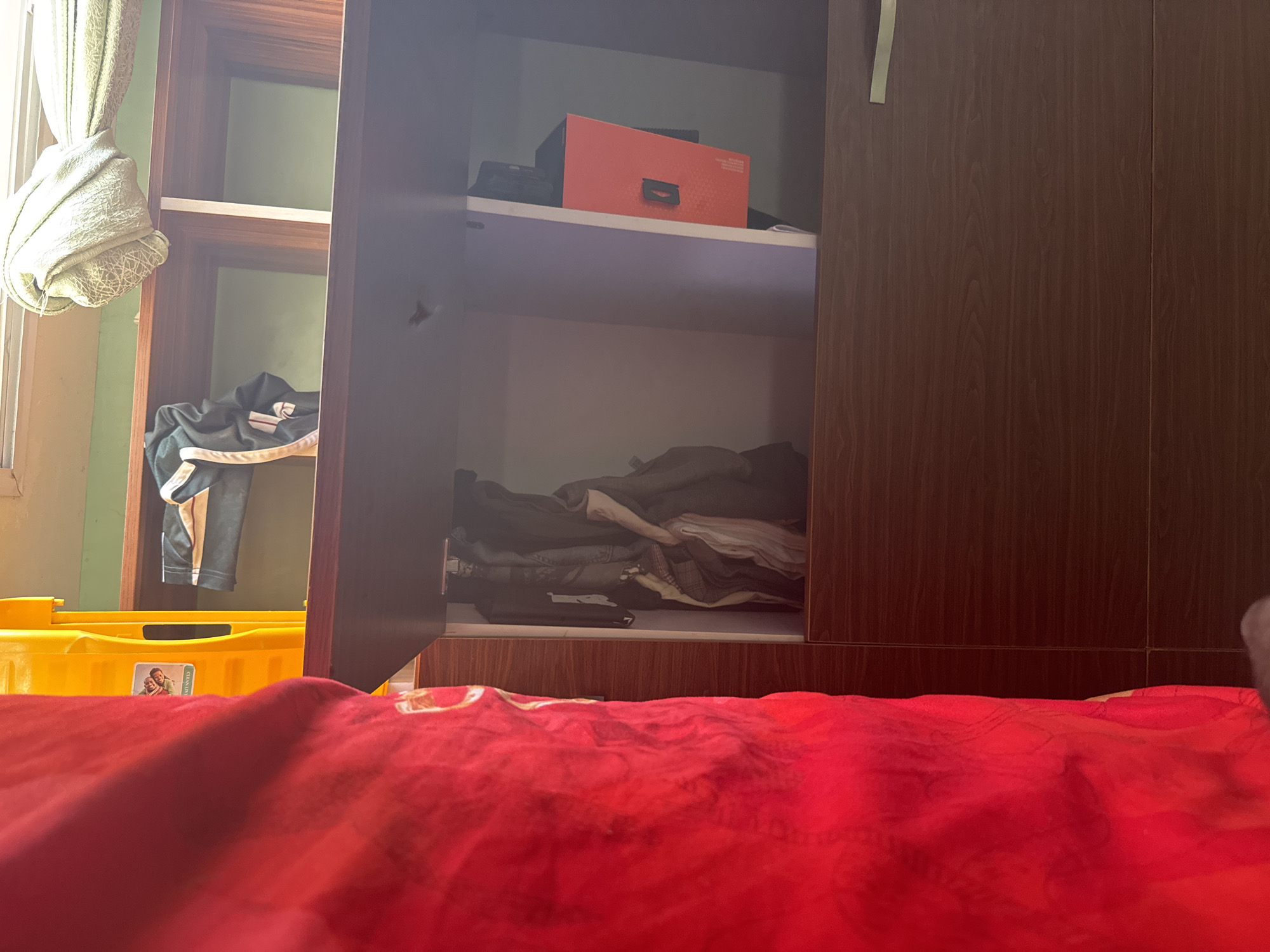 The image shows an open wardrobe with shelves full of folded clothes and some boxes. On the top shelf, there is an orange box with a black handle and some black items. The shelves below are filled with a pile of neatly folded clothes in various colors, including dark blues, grays, and whites. To the left of the wardrobe, a yellow plastic container is visible, with a sticker on it that appears to have two people depicted. A glimpse of a red blanket or bedspread fills the foreground, suggesting a bedroom setting. The wardrobe itself is made of dark wood, and the room has a light green wall visible in the background. The lighting suggests it is daytime. The overall scene is one of domestic organization and storage. No specific activity is taking place, but the presence of folded clothes implies a recent tidying or preparation for use. The location cues, such as the style of the wardrobe and the general feel of the room, are consistent with a residential setting in Narayi, Nigeria.
