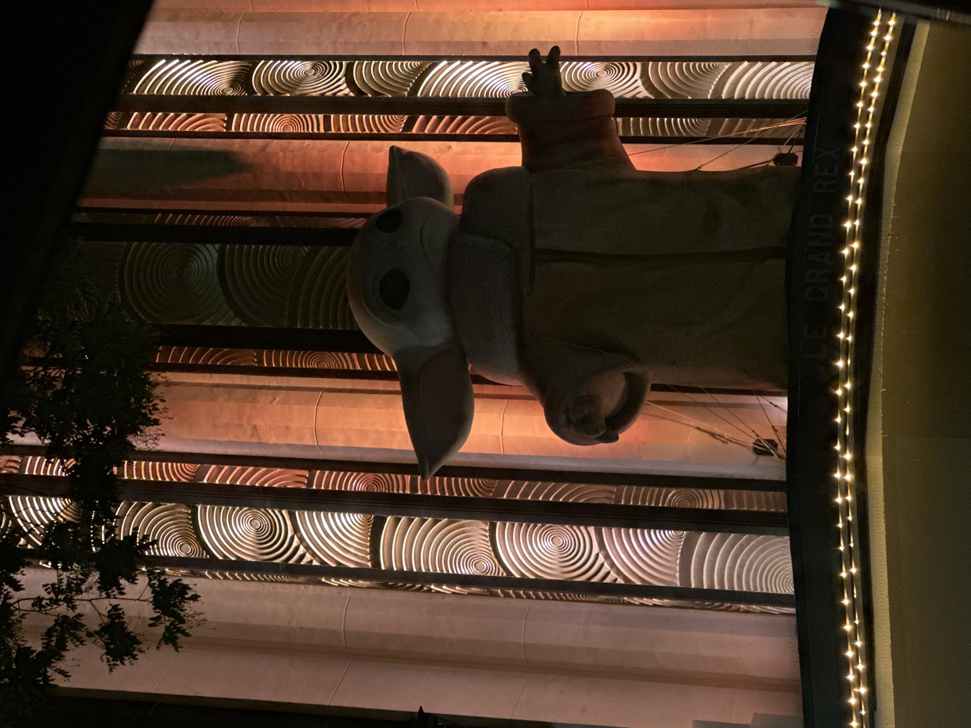 The image shows a nighttime view of a statue of Grogu (Baby Yoda) sitting atop a structure identified as "Le Grand Rex". The building exterior features a decorative design of concentric circles. String lights illuminate the right side of the frame. Part of a tree is visible on the left. The location is Paris, France.