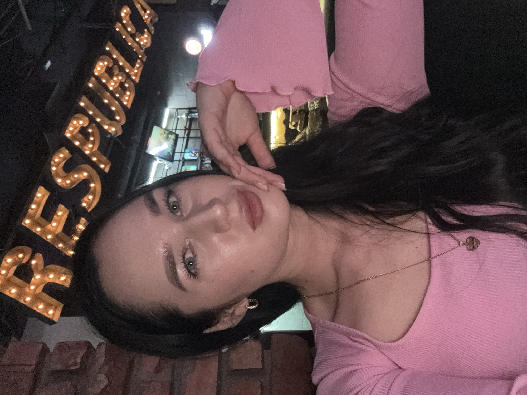 A woman with long, dark hair and light green eyes is taking a selfie. She is wearing a pink, long-sleeved shirt with ruffled cuffs and a heart-shaped pendant necklace. She has on small, hoop earrings. Her left hand is resting on her cheek. In the background, there is a brick wall and a sign with illuminated letters that spell "REPUBL" which is consistent with a sign at "Republika" club in Novosibirsk, Russia. There are also screens showing what appears to be sports or gaming content. The lighting suggests an indoor evening setting.