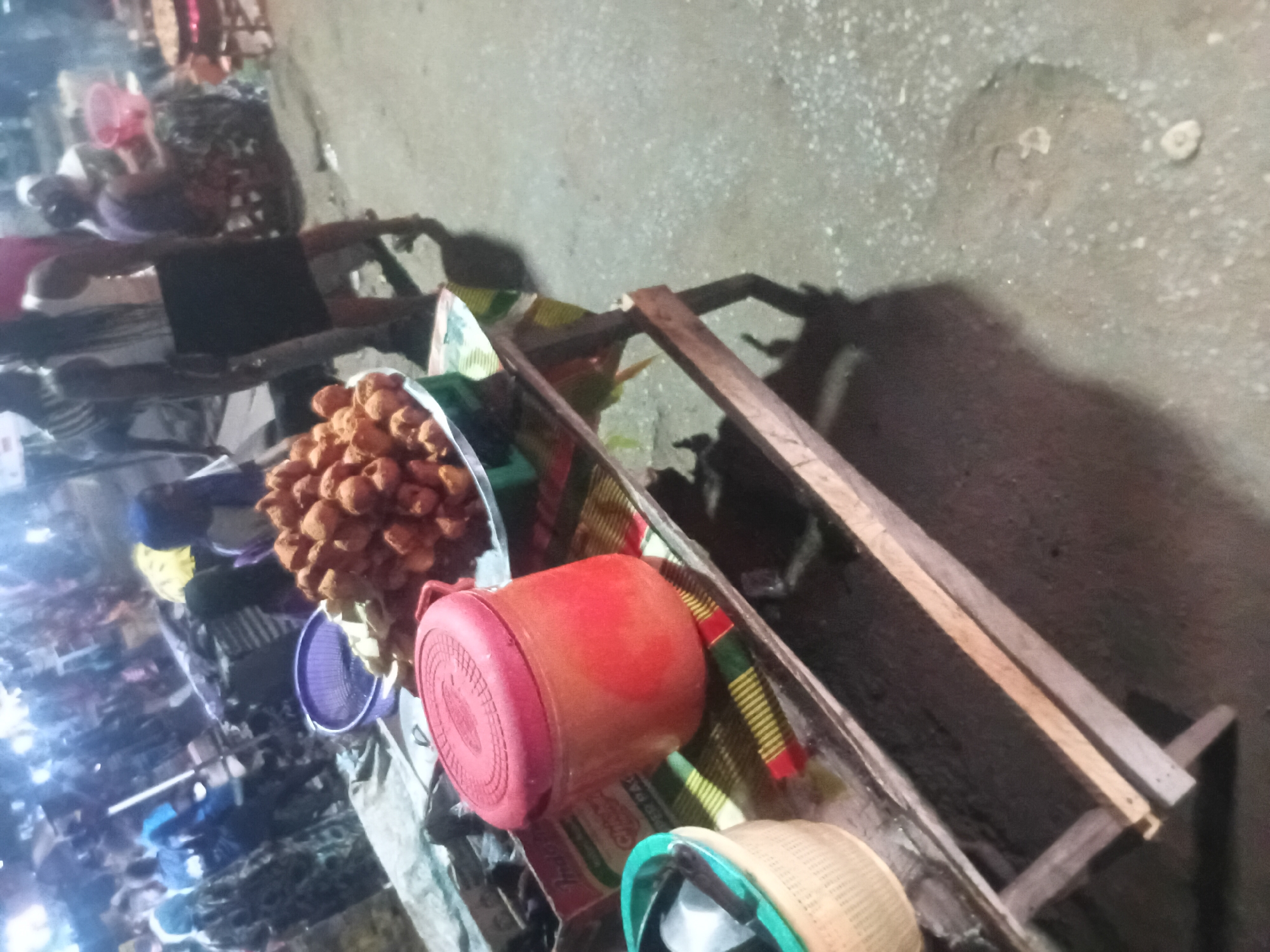 This image captures a vibrant night street food scene, likely in Lokoja, Nigeria, and is oriented on its side. In the immediate foreground, a large pile of golden-brown fried dough balls, possibly "akara" or "puff-puff," is prominently displayed in a wide, circular metal basin. Next to the food, a bright red plastic container with a lid and a purple plastic colander are visible. Further to the right, a light-colored woven basket with a distinct blue-green rim sits on a surface. These items are arranged on what appears to be a makeshift wooden stall or table, partially covered by patterned fabric.
The setting is an outdoor, bustling environment, indicative of a night market or a busy street. The ground is a rough, textured grey surface. The scene is illuminated by artificial light sources, creating significant glare and reflections, particularly in the mid and far background, suggesting a lively atmosphere. Many people are visible in the background, mostly blurred and indistinct due to the low light and movement, contributing to the sense of a crowded market. Their forms are largely silhouetted against the brighter lights. The overall ambiance points to late evening or night. No discernible text is clearly readable on any objects within the frame. The image portrays a common sight of street-side commerce and community activity after dark.