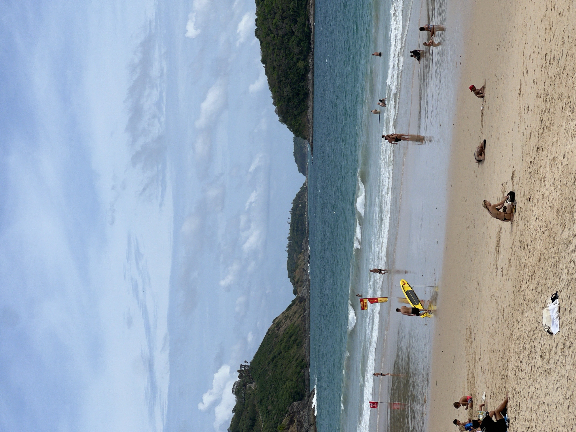 The image depicts a beach scene. The location is unknown. The beach has yellow sand and is populated with people engaging in activities such as swimming, sunbathing, and relaxing. Lifeguard flags and a yellow surfboard are visible near the shoreline. The water is blue with small waves. A green, hilly landscape borders the beach on one side, and a cloudy sky is overhead.
