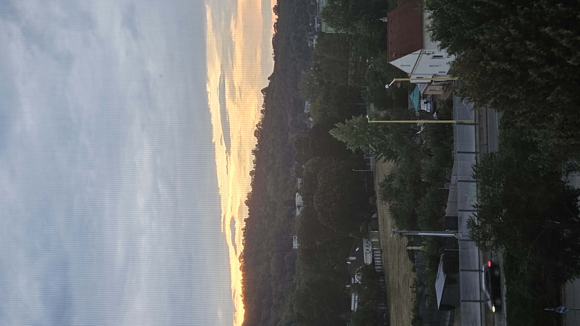 The image depicts an overcast sky with streaks of orange and yellow light visible on the horizon, suggesting either sunrise or sunset. A dark, tree-covered hill dominates the middle ground, with scattered buildings visible on its slopes and at its base. In the foreground, a road is visible with a dark-colored car driving on it, its brake lights illuminated. To the right, a white house with a red roof stands amongst trees, and a yellow lamppost is nearby. A green umbrella is visible behind the lamppost. The scene appears to be a residential area overlooking a natural landscape, likely taken from a higher vantage point. The image captures a moment of transitional light in a suburban or semi-rural setting.