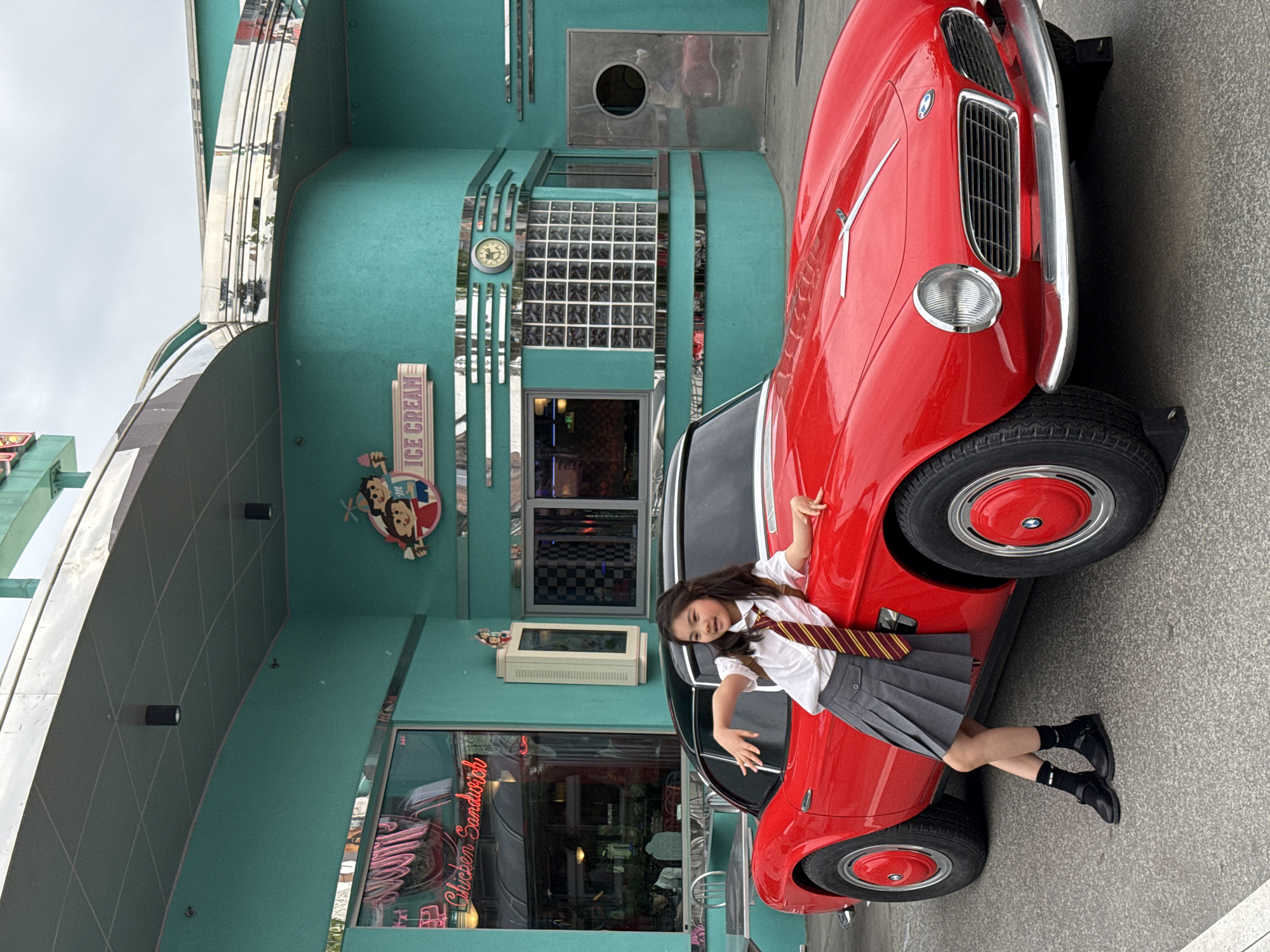 A young person wearing a white short-sleeved shirt, a gray pleated skirt, a striped tie, black socks, and black shoes poses next to a red vintage car parked outside a teal building with retro-style architecture. The building has signs reading "Ice Cream" and "Chicken Sandwich" in a stylized, neon-like font. The location is possibly a film set, tourist attraction, or themed restaurant within Tongzhou District, China.