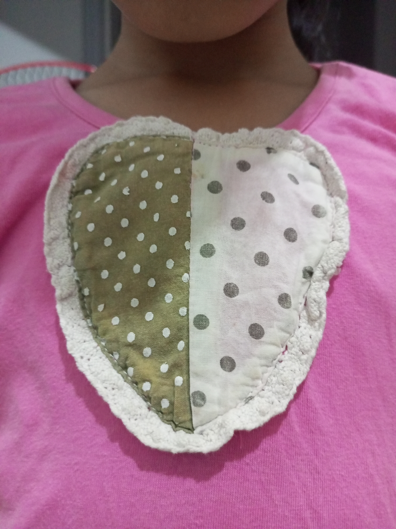 The image shows a close-up view of a pink t-shirt with a heart-shaped applique sewn onto the chest area. The heart applique is divided vertically into two halves. The left half is made of olive green fabric with white polka dots, and the right half is made of white fabric with dark grey polka dots. The heart is outlined with a white crocheted trim. The top portion of the image shows the neck and chin of a person wearing the t-shirt, but their face is not visible. The background is blurred and appears to be indoors. The image is taken from a slightly high angle, looking down at the t-shirt.
