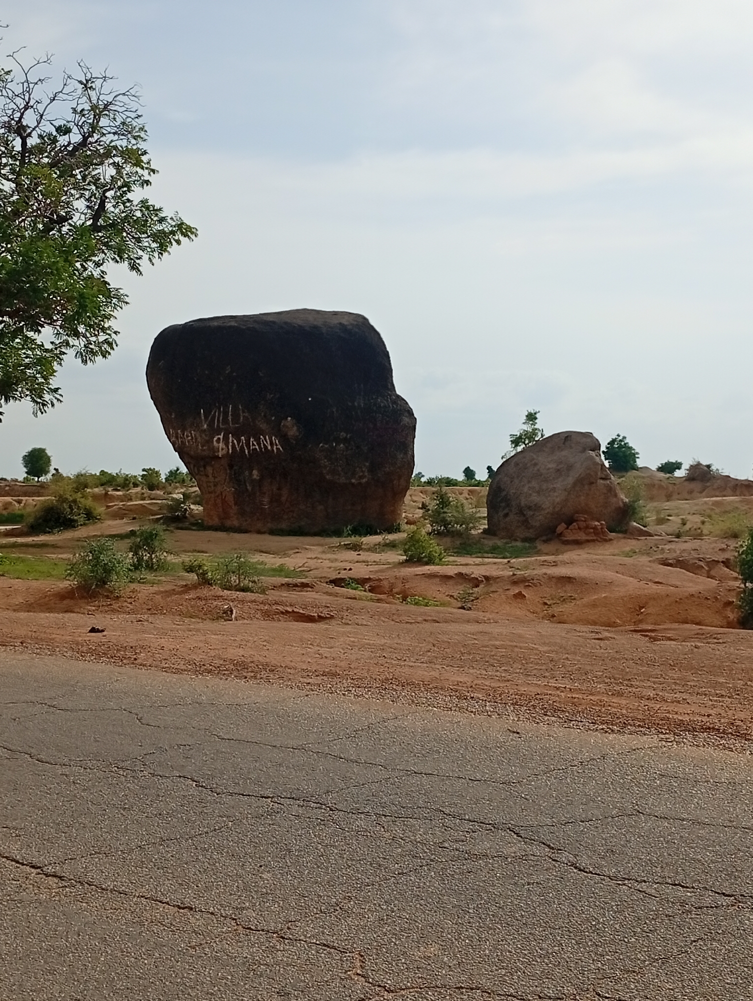 The image shows a natural landscape with two large boulders in the city of Shuwa, Nigeria. The foreground features a cracked asphalt road. In the mid-ground, there are the two boulders situated on a sandy, reddish-brown terrain with sparse vegetation. The larger boulder, on the left, is dark and imposing, with white graffiti that appears to read "VILLA" and "MANA". To its right, a smaller, lighter-colored boulder rests on the ground. The background consists of a hazy sky and a few scattered trees and bushes, suggesting a semi-arid environment. The scene is devoid of any human presence or activity.