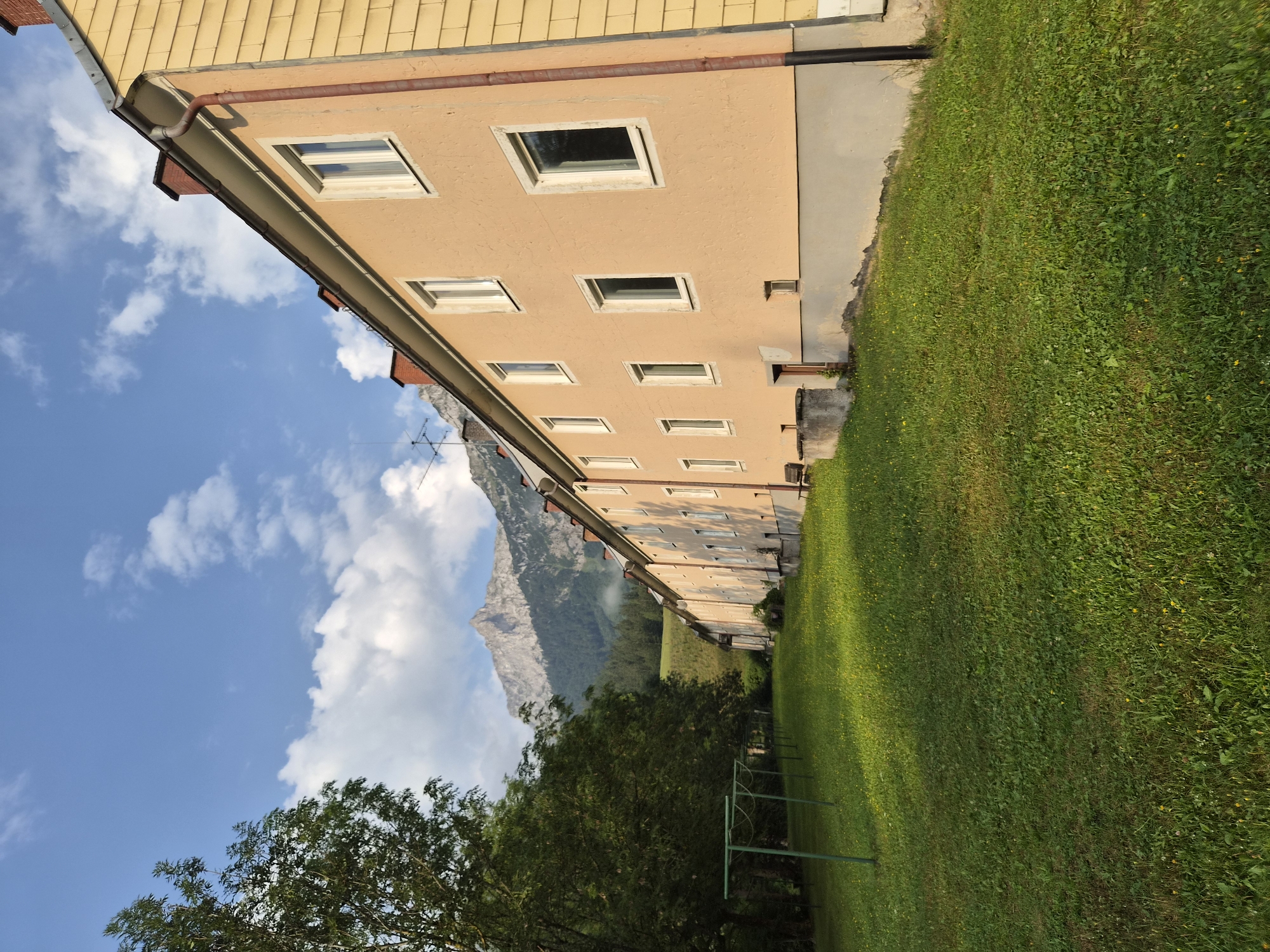 The image shows a long, three-story building with a pale orange facade and yellow siding on the right side, situated in a grassy area. The building has numerous windows, a red-brick chimney, and a red gutter system. In the background, a large, rugged mountain range is visible, with trees dotting the lower slopes and a cloudy blue sky above. In the foreground, a lush green lawn is mowed, with some small yellow flowers scattered throughout. A green metal clothesline rack stands on the left side of the lawn. The scene appears to be in a rural or semi-rural setting, likely in Eisenerz, Austria, given the mountainous terrain. There are no people or visible actions depicted in the image.