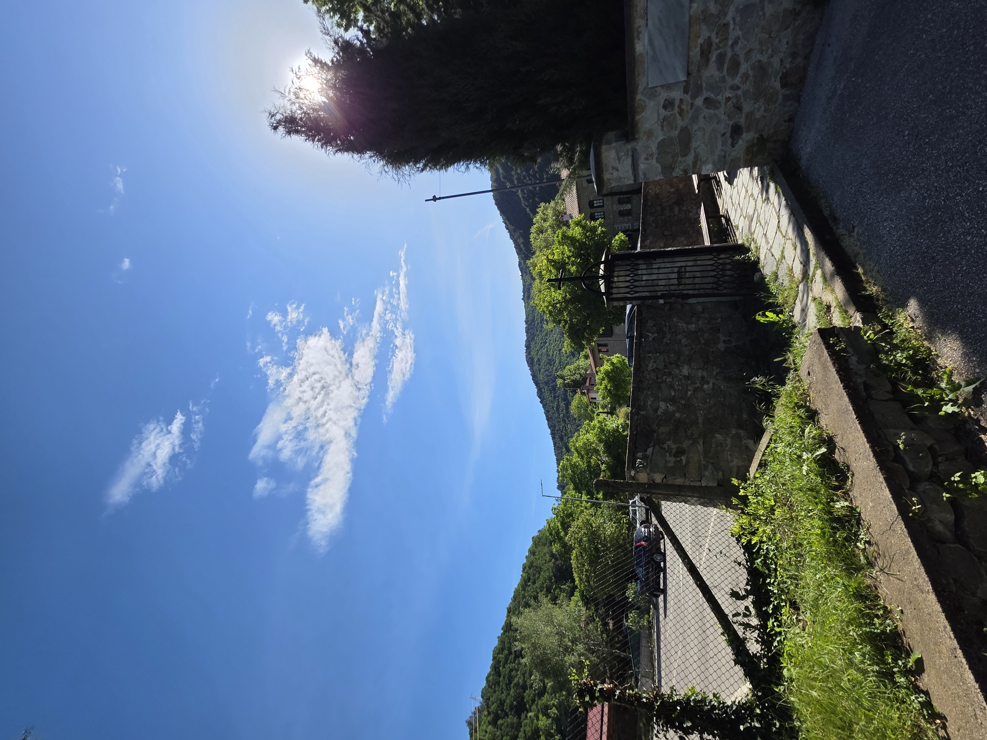 The image captures a scene in Bachkovo, Bulgaria. The viewpoint is from a slightly elevated position looking towards a mountainside densely covered with green trees. A section of the sky is visible, blue with scattered white clouds, and the sun is visible through the trees. In the foreground, there is a stone wall and a paved area with vegetation growing alongside it. A road runs parallel to the wall. A portion of a building, likely a house, is visible with a decorative iron gate. A utility pole is observed in the middle ground.