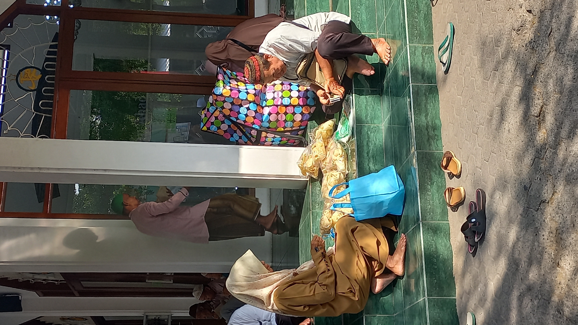 In Kenayan, Indonesia, an outdoor scene unfolds on the green tiled steps of a building, likely a place of worship given the attire of the individuals. Two people are seated on the steps, engaged in what appears to be a transaction or the distribution of goods. A woman, wearing a tan hijab and matching long-sleeved garment, sits on the left, looking down at something in her hands, possibly food items arranged in plastic bags. A blue tote bag rests beside her. To her right, a man in a white long-sleeved shirt and dark trousers, wearing a red and black patterned cap, is crouched, also looking at items in his hands, which seem to be small packets. He has a bag or satchel resting by his side.
In the background, a man in a light pink tunic and a brown sarong walks away from the camera, holding a light-colored object. The architecture features large wooden-framed glass doors and white pillars, with a stained-glass design above the doors. A large, colorful wheeled bag adorned with polka dots and the word "LOVE" is situated near the man in the white shirt. The ground in front of the steps is paved with hexagonal tiles, where several pairs of sandals are scattered: a pair of black slide sandals, two pairs of brown sandals, and a single green and white flip-flop. The scene is bathed in bright sunlight, casting shadows on the paved area.