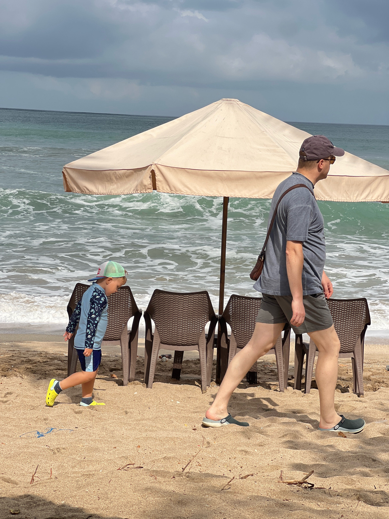 A full-shot image captures a beach scene in Kuta, Indonesia, under a cloudy sky. The ocean, with its white-capped waves, occupies the upper half of the frame. In the foreground, a man and a young child walk on the sand. The man, wearing a grey t-shirt, grey shorts, a baseball cap, and sunglasses, has a brown satchel slung across his chest. The child, in a blue long-sleeved rash guard, dark blue shorts, and a green and white baseball cap, walks ahead of the man. Two empty brown plastic beach chairs are positioned behind the child, and a large beige beach umbrella stands to the left of the man, partially obscuring the ocean. The sandy beach is sparsely scattered with small pieces of driftwood.