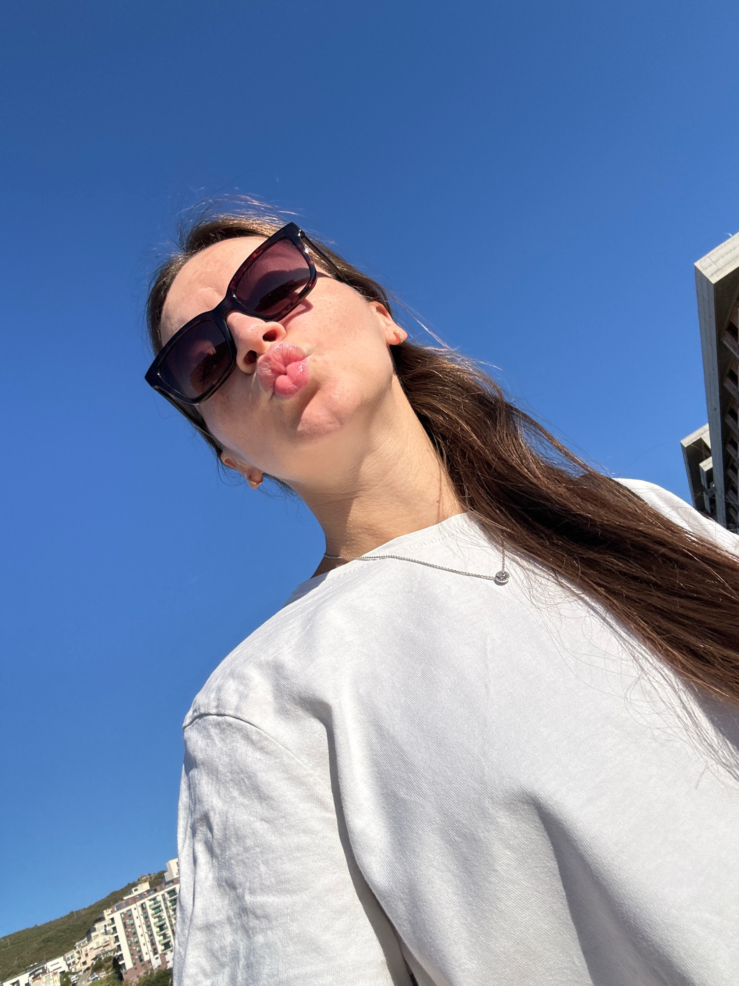A young woman with long brown hair, wearing sunglasses and a white t-shirt, poses for a selfie outdoors. She has her lips pursed for a kiss. The background is a clear blue sky, with a modern building visible on the right and buildings on a hillside to the left, suggesting an urban or semi-urban environment, possibly along the coast. The lighting indicates it is daytime.
