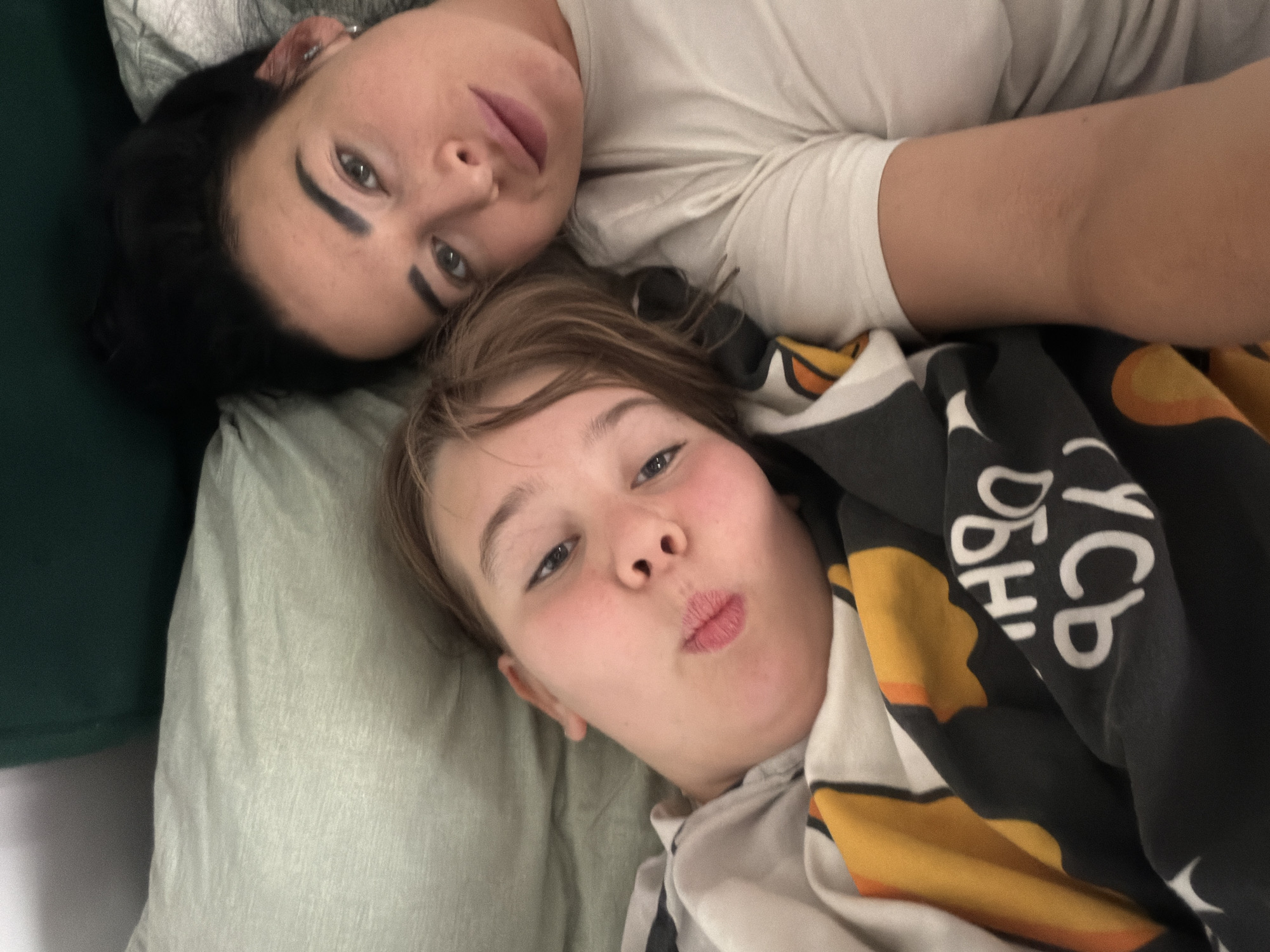 The image shows two individuals, presumably a woman and a child, lying down next to each other. The woman has dark hair and is wearing a light-colored shirt. The child is partially covered with a patterned blanket. They are lying on a green surface, possibly a bed or couch. The background is out of focus, making it difficult to ascertain the exact location. No information suggests it is within Veliky Novgorod, Russia.