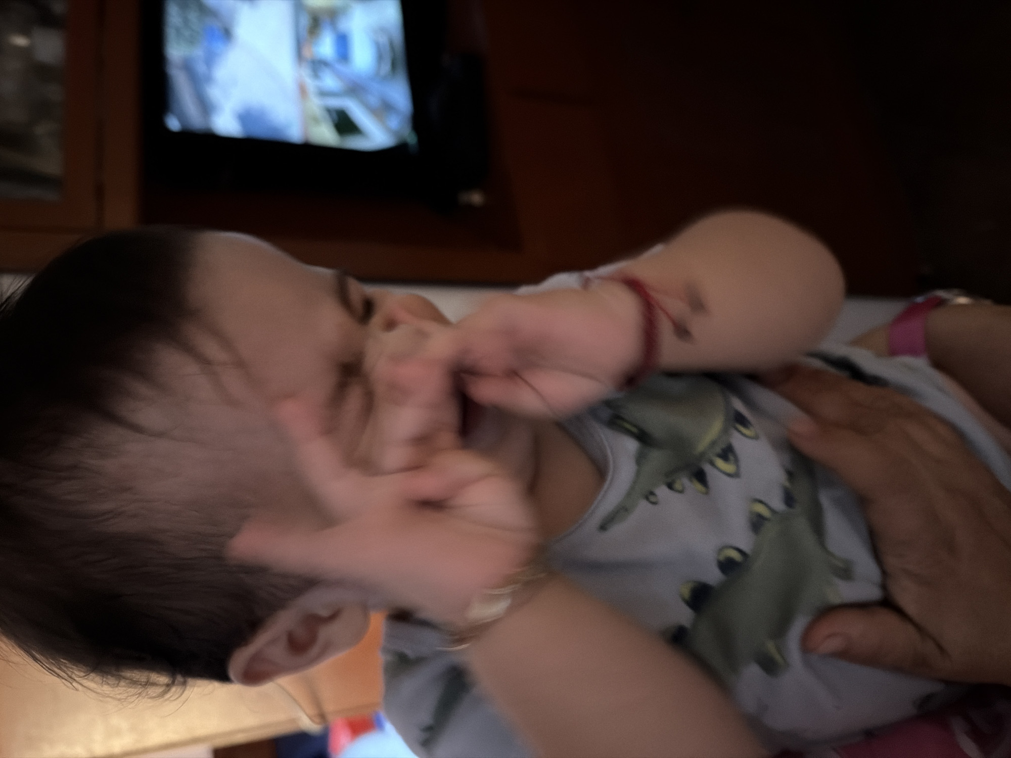 A baby wearing a dinosaur-printed onesie is lying down with a hand near their face. An arm is holding the baby. A television is in the background. The image was taken indoors. There is no indication of this image being in Reforma, Mexico.