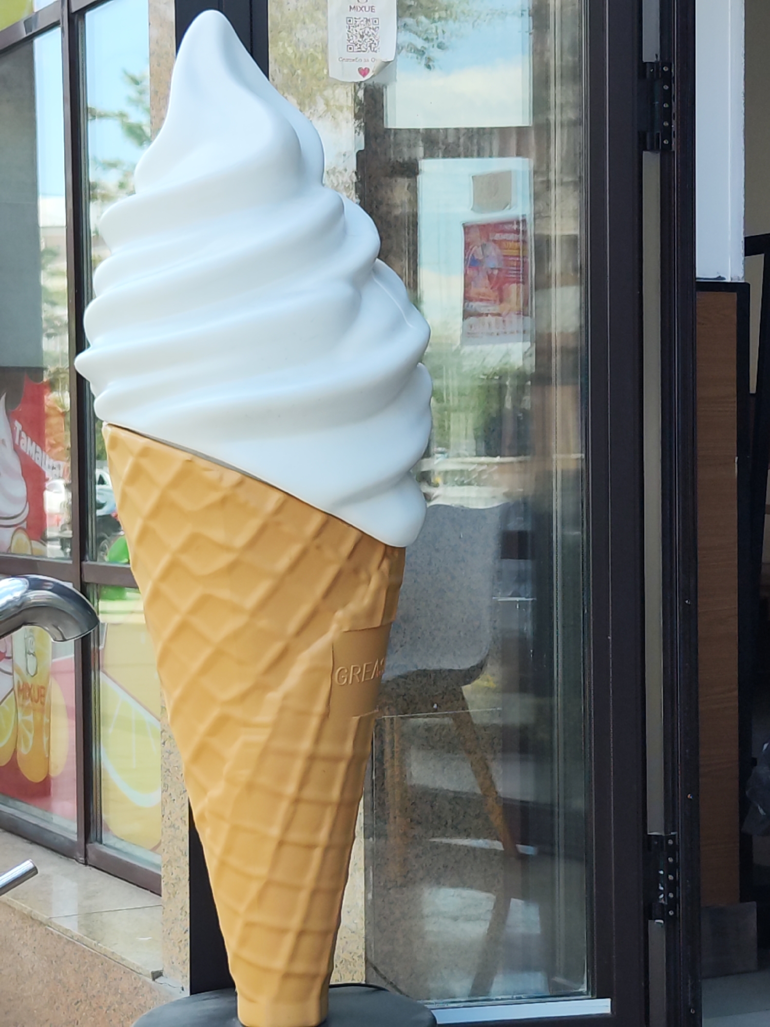 A large, white soft-serve ice cream cone statue stands in front of a glass storefront. The statue is made of plastic and has a realistic waffle cone base. The storefront appears to be a cafe or ice cream shop, with a window displaying advertisements for soft-serve ice cream. The background shows a street scene in Astana, Kazakhstan, with trees and buildings visible. The weather appears to be sunny.