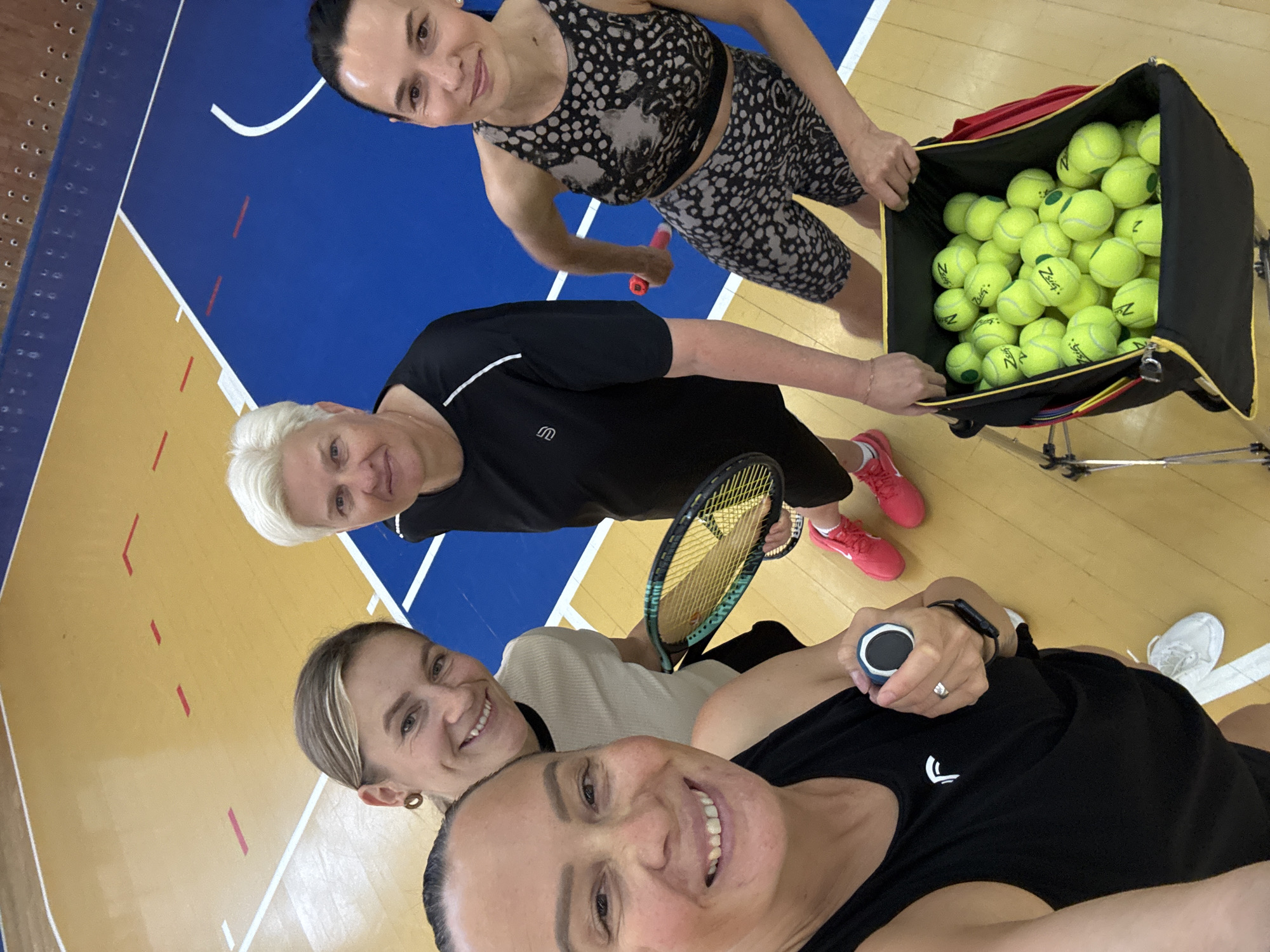 Four women are posing for a selfie in a gymnasium. One woman at the bottom of the frame is holding the camera. Two women are in the middle of the frame, with one holding a tennis racket. The last woman is standing behind a basket full of tennis balls. The gymnasium floor is marked with lines for basketball and volleyball, and the walls are blue and yellow. The location is Chaikovsky, Russia.