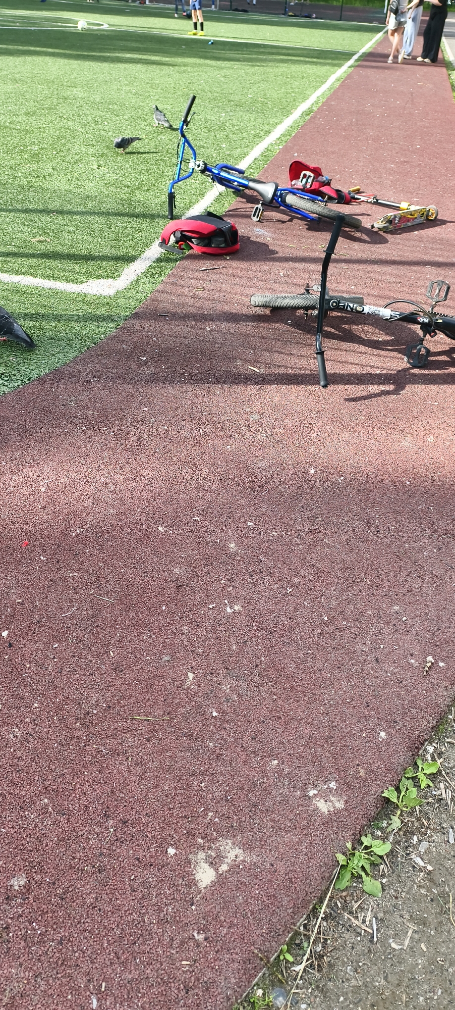 The image captures an outdoor sports complex, featuring an artificial green grass field and an adjacent reddish-brown, granular rubberized track. In the midground, on the track, a blue BMX-style bicycle lies on its side, next to a red and black helmet and a yellow and black scooter. In the foreground, part of a black and white bicycle is visible, also lying on the track, with the text "ONFO" visible upside down on its tire.
Two pigeons are present on the artificial grass field, near the white boundary line that separates the track from the field. In the background, on the grass field, several people are visible. One person in a blue top and yellow socks appears to be involved in an activity, possibly soccer, as a white ball is also seen on the field. Other figures are partially visible further back on the track.
The scene is brightly lit by daylight, casting distinct shadows from the bicycles and other objects onto the track, indicating a clear, sunny day. The location cues suggest a public or school sports facility.