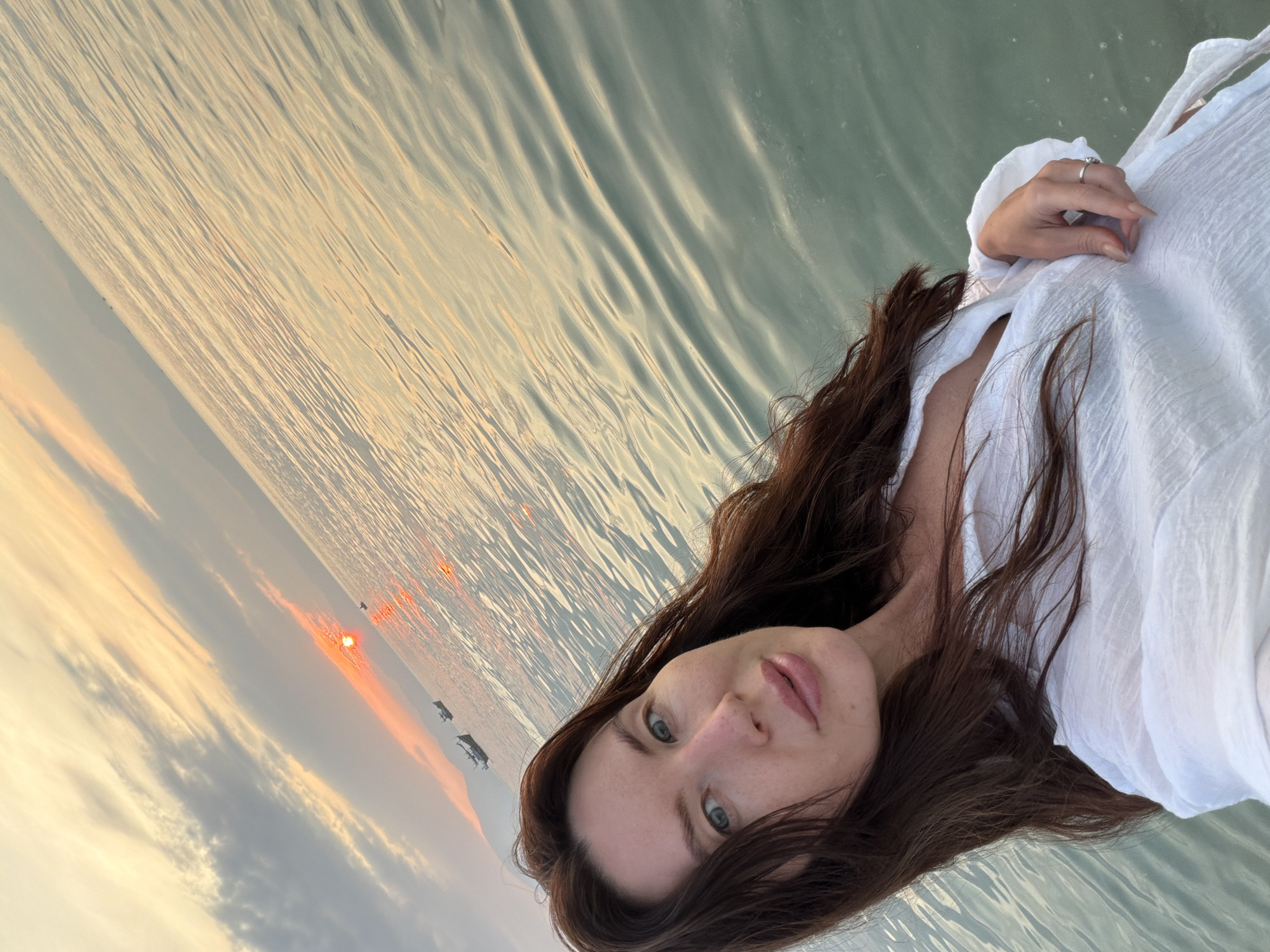 A woman is captured in a selfie at sea during sunset. She has long, dark brown hair and is wearing a white, loosely-buttoned shirt. The water is calm with ripples reflecting the warm hues of the setting sun. In the background, two boats are visible on the horizon, and a faint outline of landmasses can be seen. The overall scene suggests a serene and picturesque moment. The image was taken in the vicinity of Dong Ninh Hoa Ward, Vietnam.