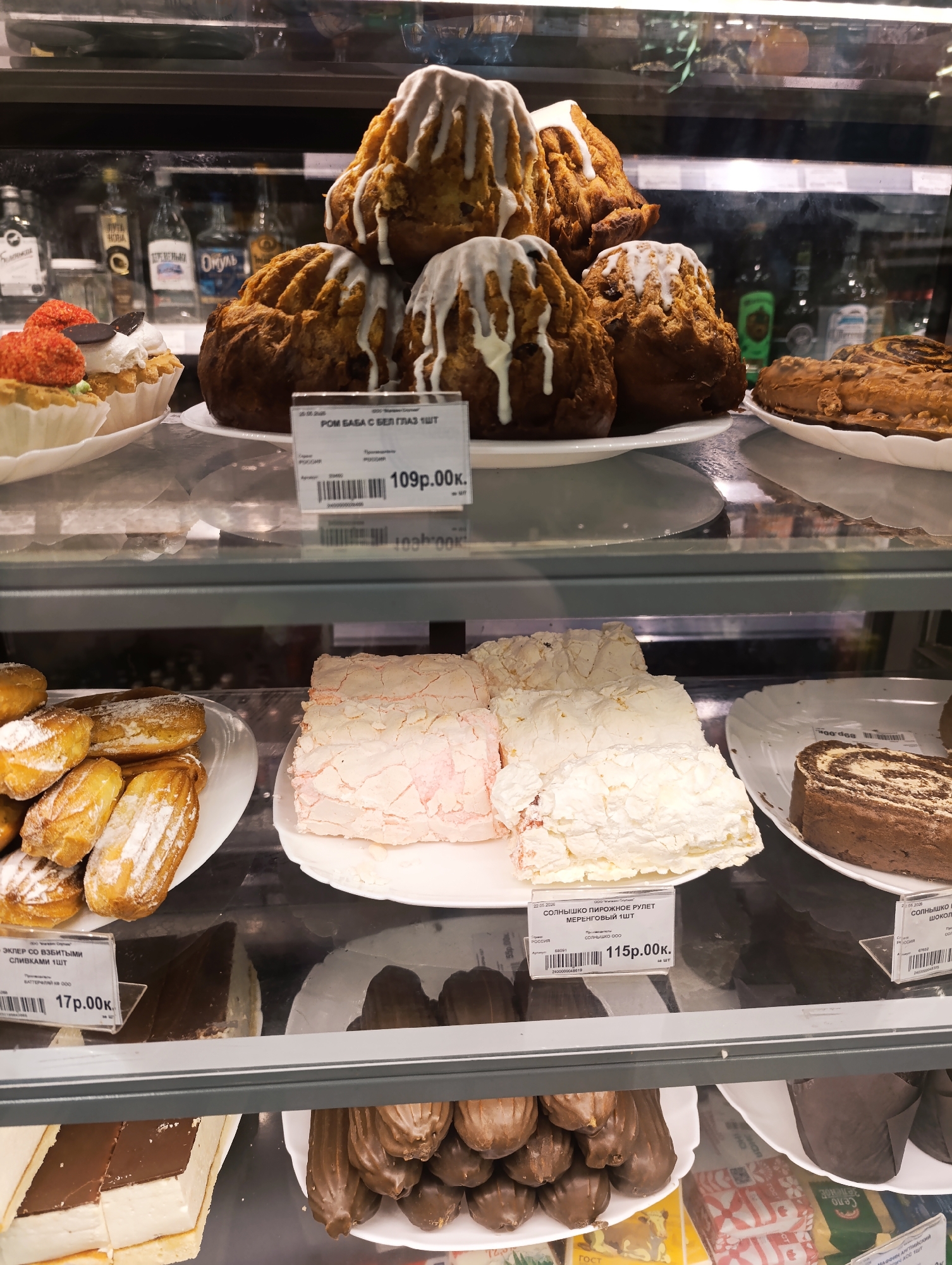 This image shows a display case filled with pastries. On the top shelf, there are rum babas with white glaze piled high on a plate, as well as other assorted cakes and tarts. The middle shelf contains eclairs dusted with powdered sugar, meringue rolls, and chocolate-swirl rolls. The bottom shelf is filled with an assortment of cakes and chocolate-covered eclairs. Each item has a price tag in Russian, indicating the price in rubles and kopeks. The location is a pastry shop or bakery, judging by the display of various baked goods. The location is Unknown, Unknown.