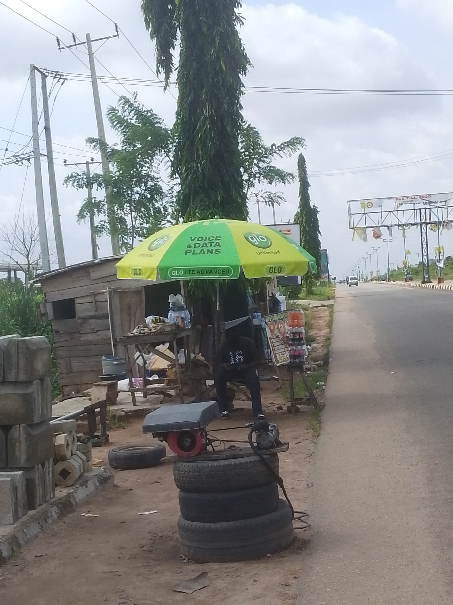 This image depicts a roadside scene in Abeere, Nigeria, likely near a major highway. In the foreground, to the left, a stack of grey concrete blocks is piled on a dirt surface. To the right of the blocks, a small, weathered wooden shack is visible, with a table set up in front of it displaying various items, including bottled water and what appear to be electronic accessories. A green and yellow umbrella with "GLO VOICE & DATA PLANS" printed on it provides shade over the area.
A man, wearing a dark shirt with the number "16" on the back, is seated on a dark chair to the left of the shack, looking at his phone. Next to the shack, a stack of four car tires is arranged vertically, with a small red generator resting on the top tire, connected to a hose. The road is visible to the right of the scene, with a white car driving in the distance. Overhead, power lines and poles run along the side of the road, and tall trees are interspersed with other vegetation. Further in the background, large billboards are mounted on structures above the road. The sky is overcast with some patches of blue.