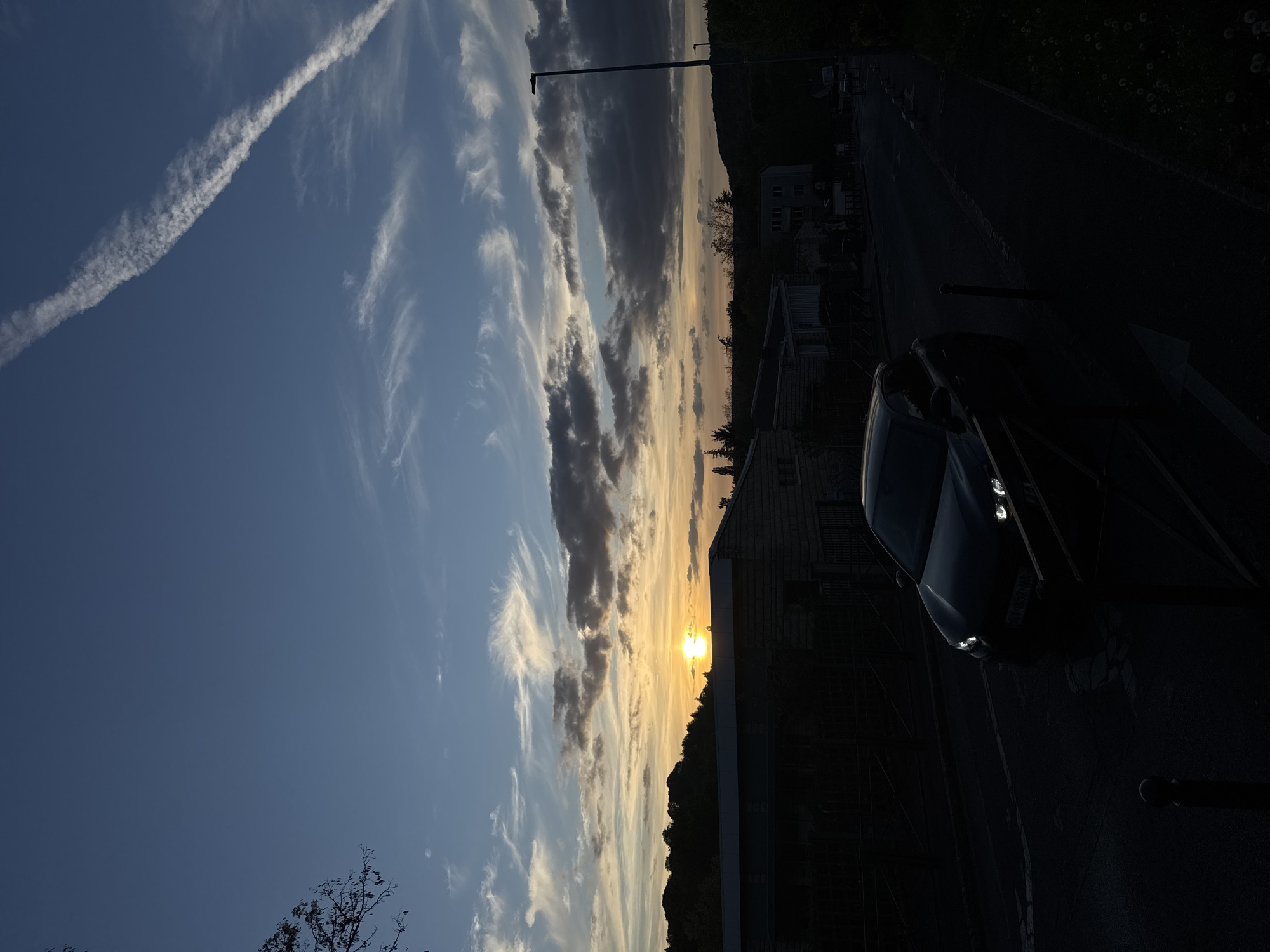 The image captures a tranquil sunset scene in Bures-sur-Yvette, France, presented in a portrait orientation as if looking sideways across a landscape. The sun is low on the horizon, glowing bright orange and yellow, partially obscured by a dark, hilly landscape that forms the skyline.
The sky exhibits a gradient of colors, transitioning from a deep blue in the upper left to lighter blues, oranges, and yellows near the horizon, indicative of either sunrise or sunset. Scattered white and grey clouds streak across the sky, with a prominent, distinct white airplane contrail cutting diagonally across the upper left quadrant.
In the foreground and midground on the right side, a dark-colored car is parked on a paved road, with its headlights faintly illuminated. Several buildings with visible architectural details like roofs and windows are visible along the street, mostly appearing as dark silhouettes against the brighter sky. A tall, slender lamppost stands vertically near the center of the frame, extending into the sky. Dark trees and foliage are visible on the distant hills and around the buildings.
The scene conveys a peaceful and serene atmosphere at the end (or beginning) of the day. No people are visible. Some text is present on the car's license plate but is unreadable due to the darkness and image resolution.